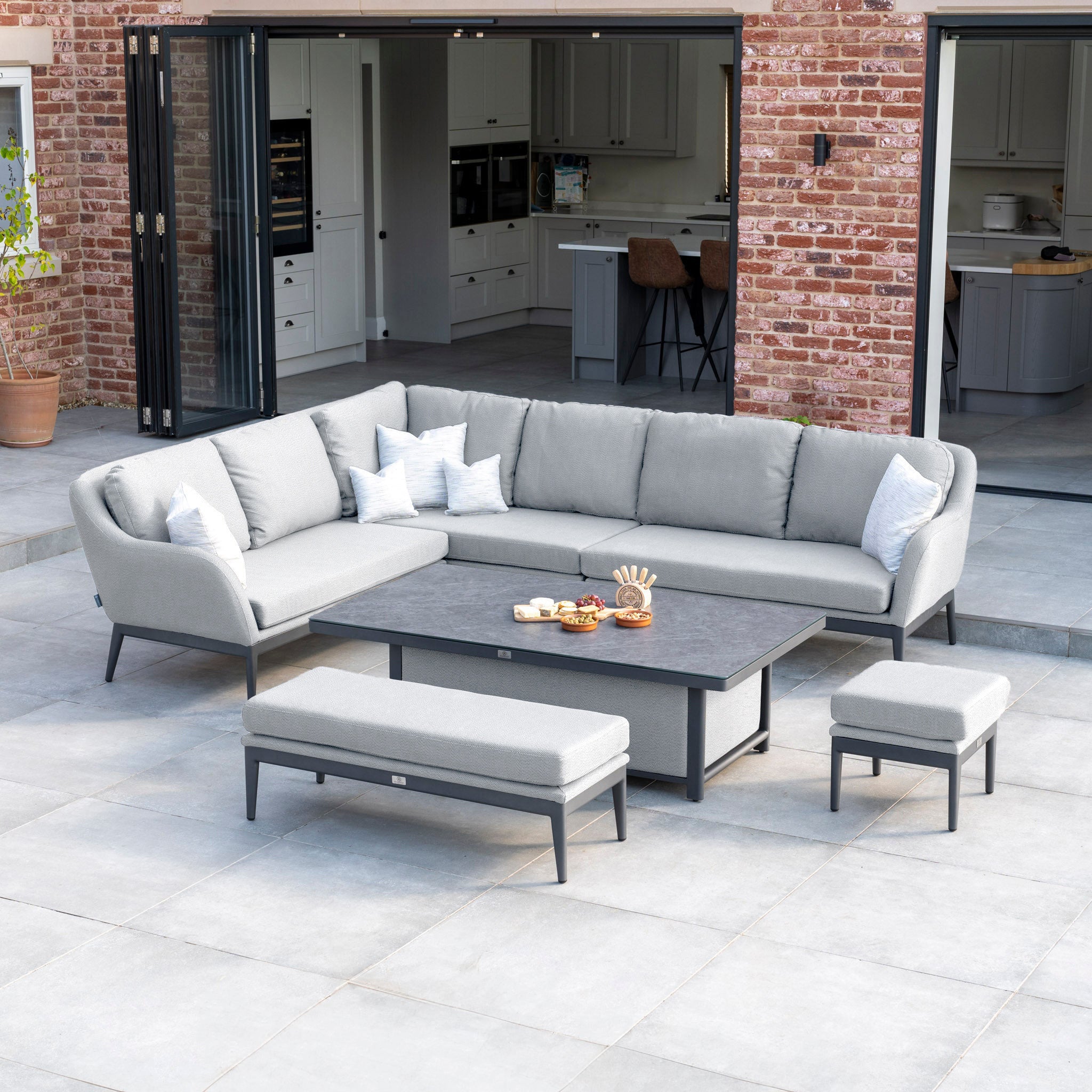 Modern outdoor patio featuring the Luna Outdoor Fabric Rectangular Corner Dining Set with Rising Table in Oyster Grey (Left Hand), complete with a matching bench and ottoman on a large tiled floor. The Luna outdoor fabric ensures the seating is maintenance-free.