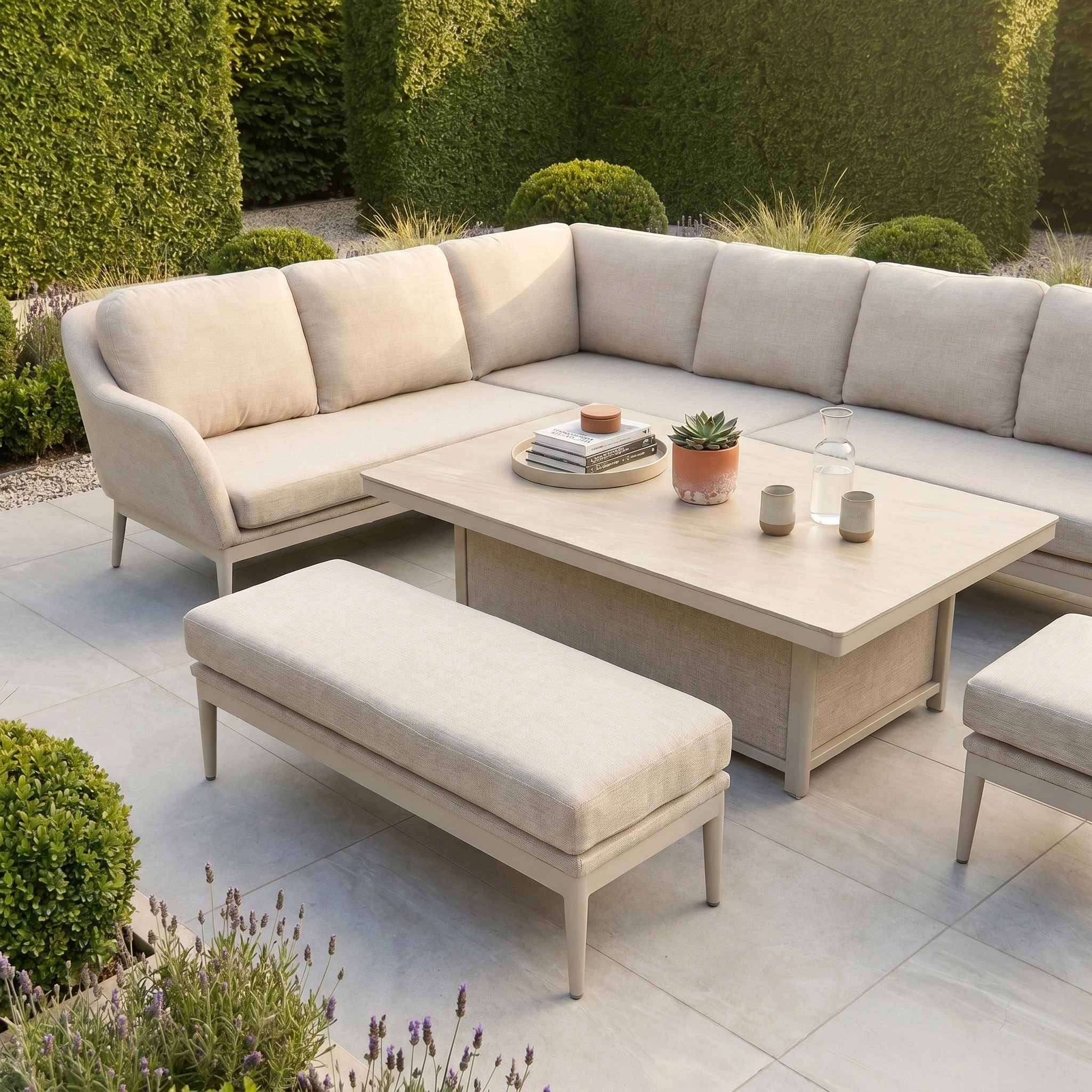 The Luna Outdoor Fabric Rectangular Corner Dining Set with Rising Table in Fawn (Left Hand) features a beige sectional, bench, and table made with all-weather materials, set on a tiled patio amid lush green bushes and plants.