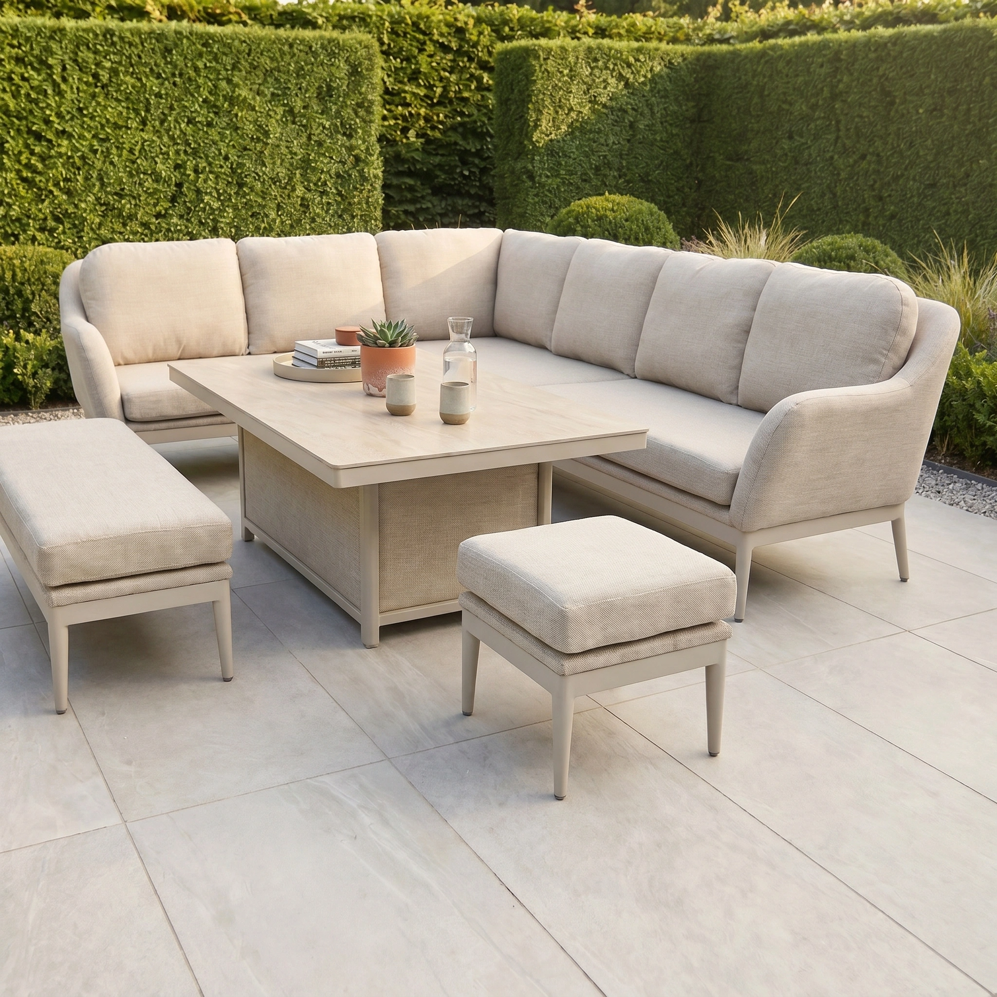 The Luna Outdoor Fabric Rectangular Corner Dining Set with Rising Table in Fawn (Left Hand) features a sectional sofa, bench, and ottoman made from all-weather material, shown on a tiled patio with green hedges.