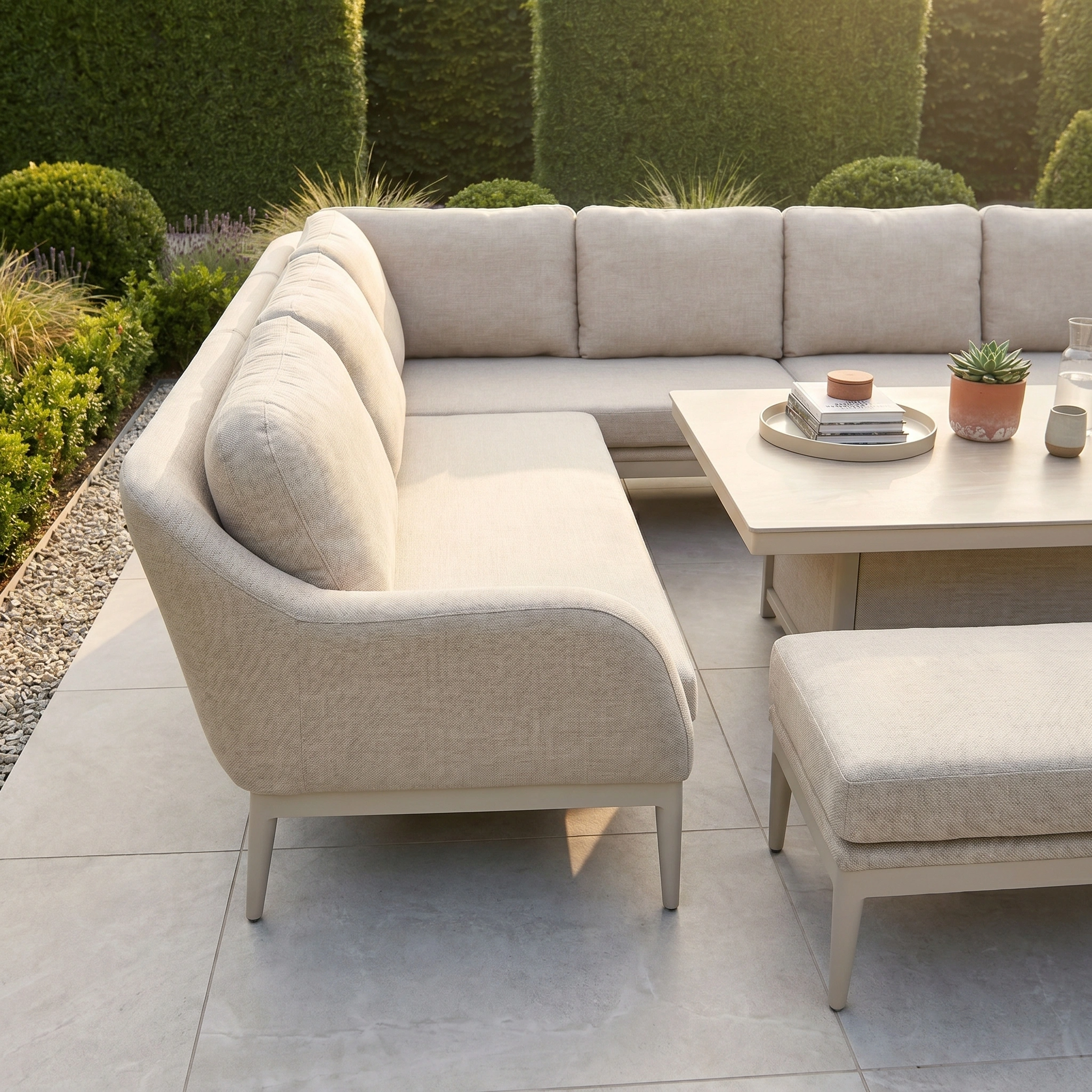 The Luna Outdoor Fabric Rectangular Corner Dining Set with Rising Table in Fawn (Left Hand) sits on a tiled patio amid lush greenery, crafted from all-weather material for lasting outdoor comfort.