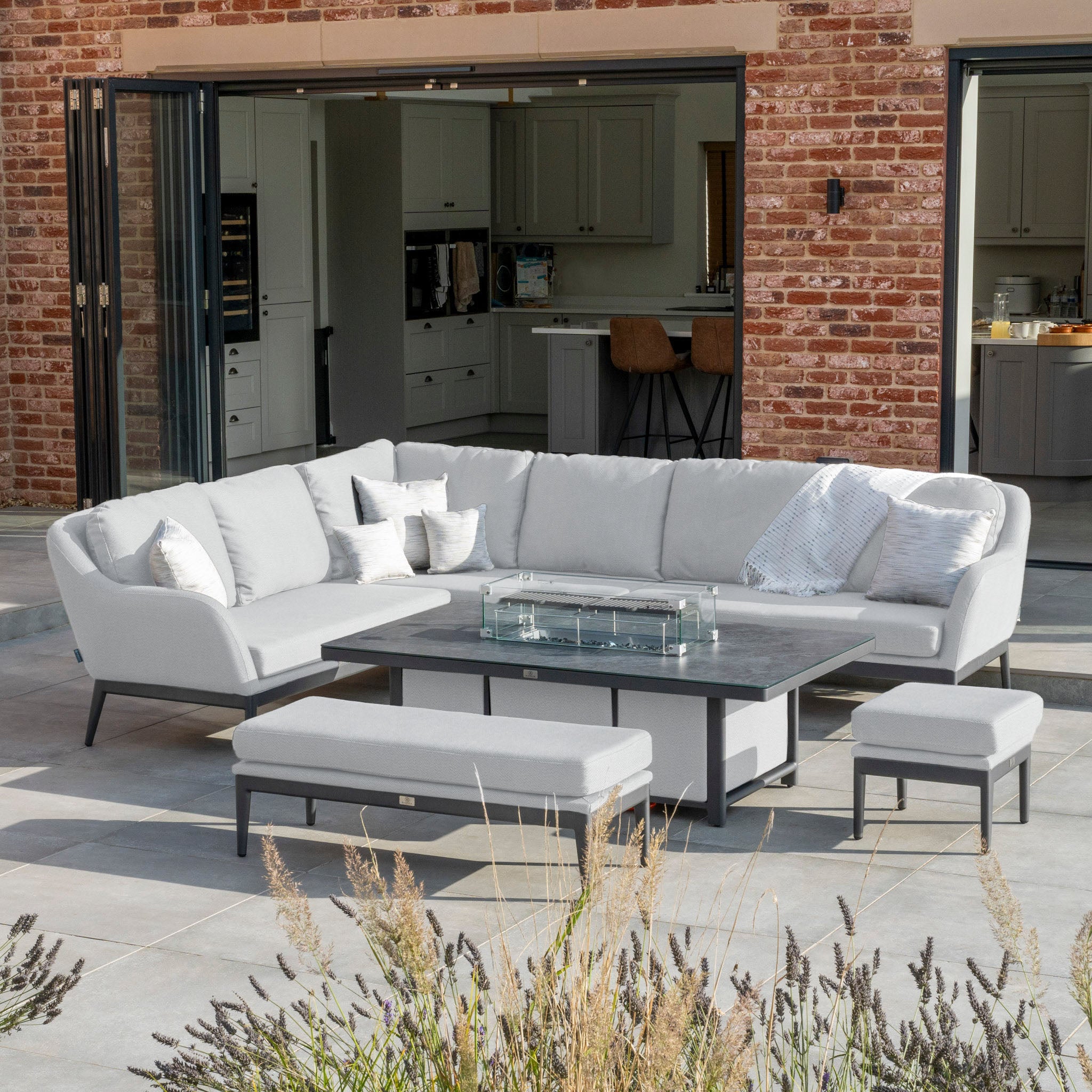 Outdoor patio with a Luna Outdoor Fabric Rectangular Corner Dining Set in Oyster Grey, featuring a maintenance-free white sectional sofa, rising firepit table, two stools, and an open view into the house kitchen.
