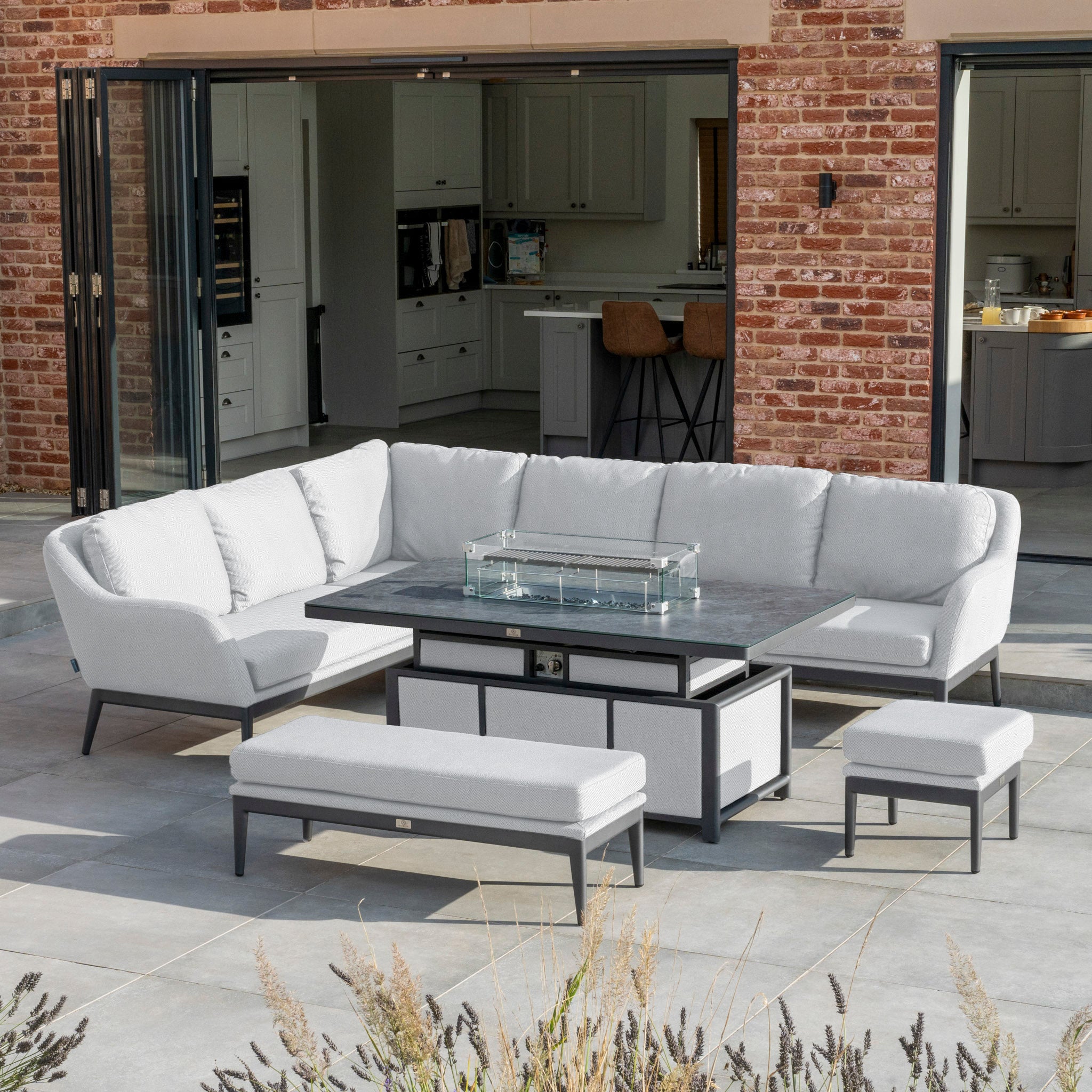 Modern outdoor patio featuring the Luna Outdoor Fabric Rectangular Corner Dining Set with Rising Firepit Table in Oyster Grey (Left Hand), complemented by a bench and stools. The setup is against a brick house with open doors.