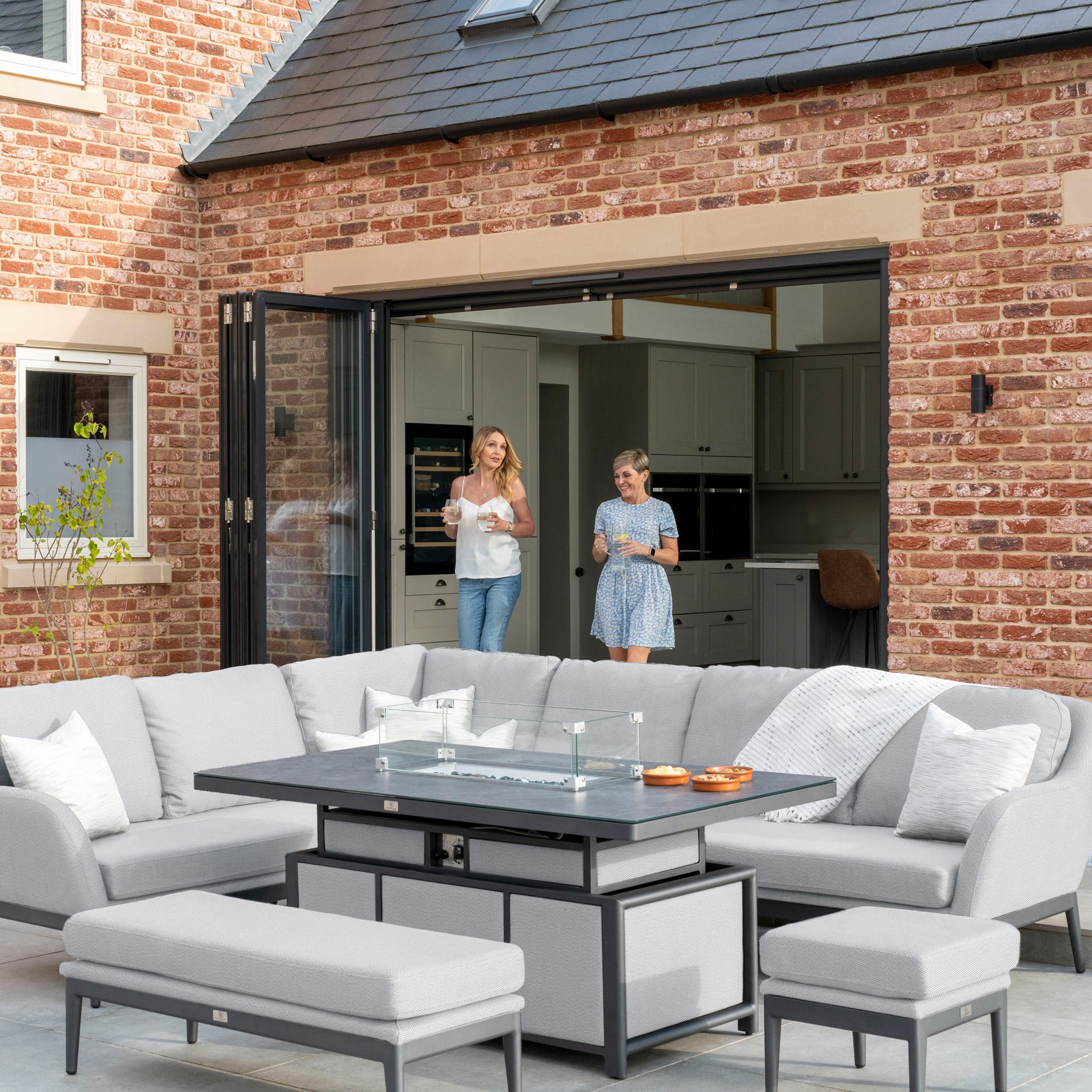 Two people standing near a house with a large patio, featuring a maintenance-free Luna Outdoor Fabric Rectangular Corner Dining Set with Rising Firepit Table in Oyster Grey (Left Hand).