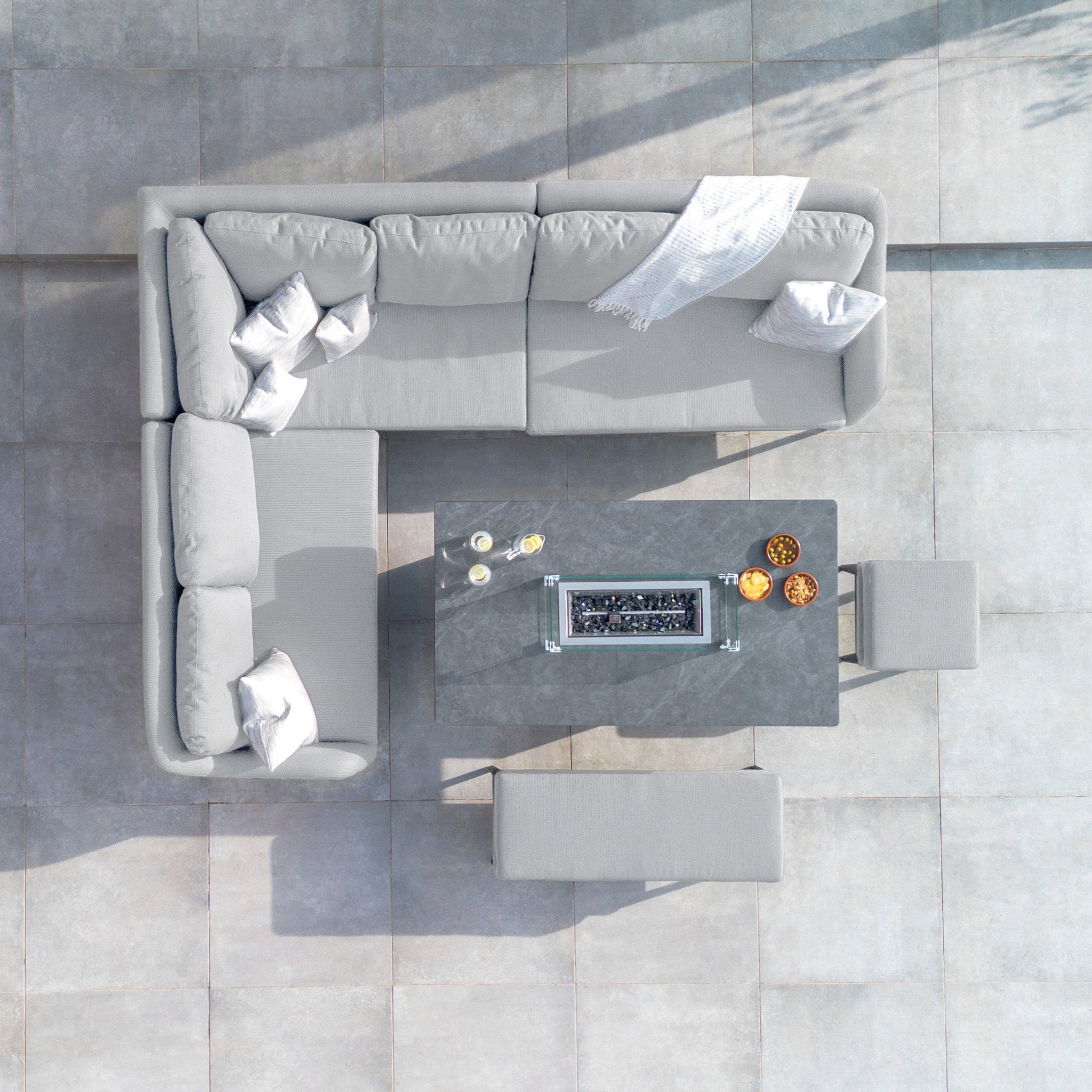 Top-down view of a patio featuring the Luna Outdoor Fabric Rectangular Corner Dining Set with a rising firepit table in Oyster Grey (Left Hand), including a grey L-shaped sofa, the table adorned with snacks, and two benches on a tiled floor.