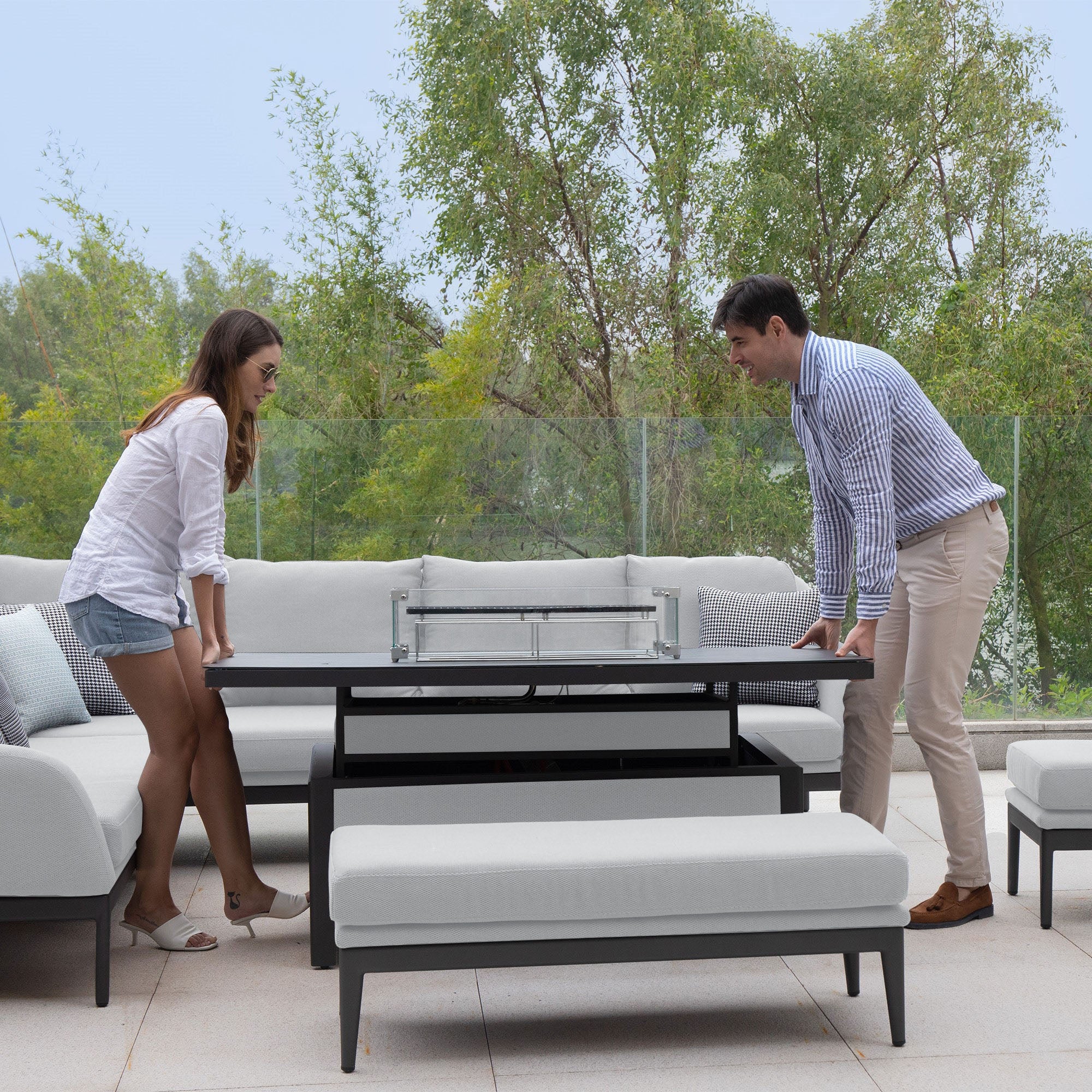 Two people lifting the maintenance-free **Luna Outdoor Fabric Rectangular Corner Dining Set with Rising Firepit Table in Oyster Grey (Left Hand)** in an outdoor seating area with a glass fence and trees in the background.