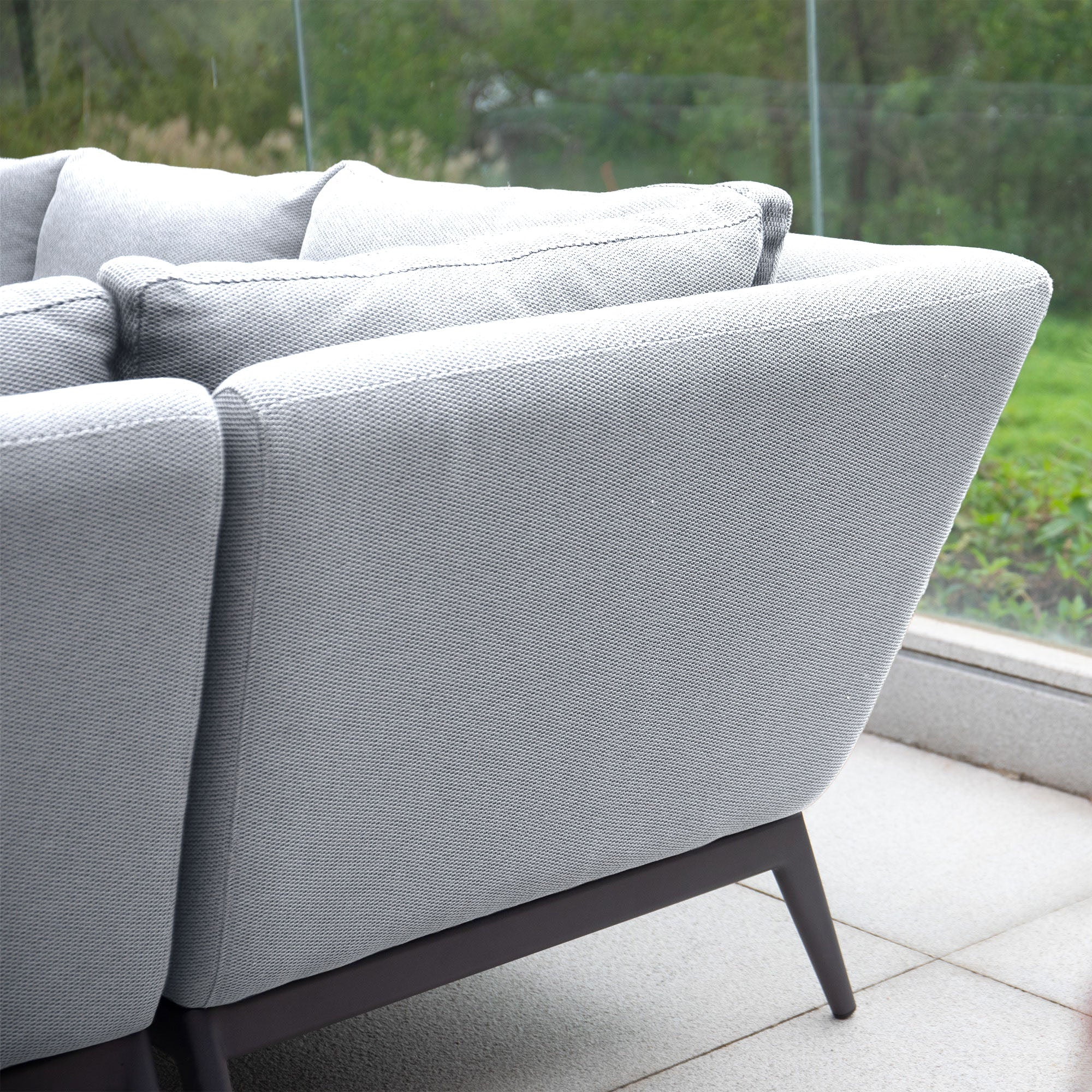 Close-up of the back of a grey Luna Outdoor Fabric Rectangular Corner Dining Set with cushions, positioned near a glass window overlooking greenery outside. Pair this maintenance-free seating with its stylish rising firepit table for the perfect outdoor escape.