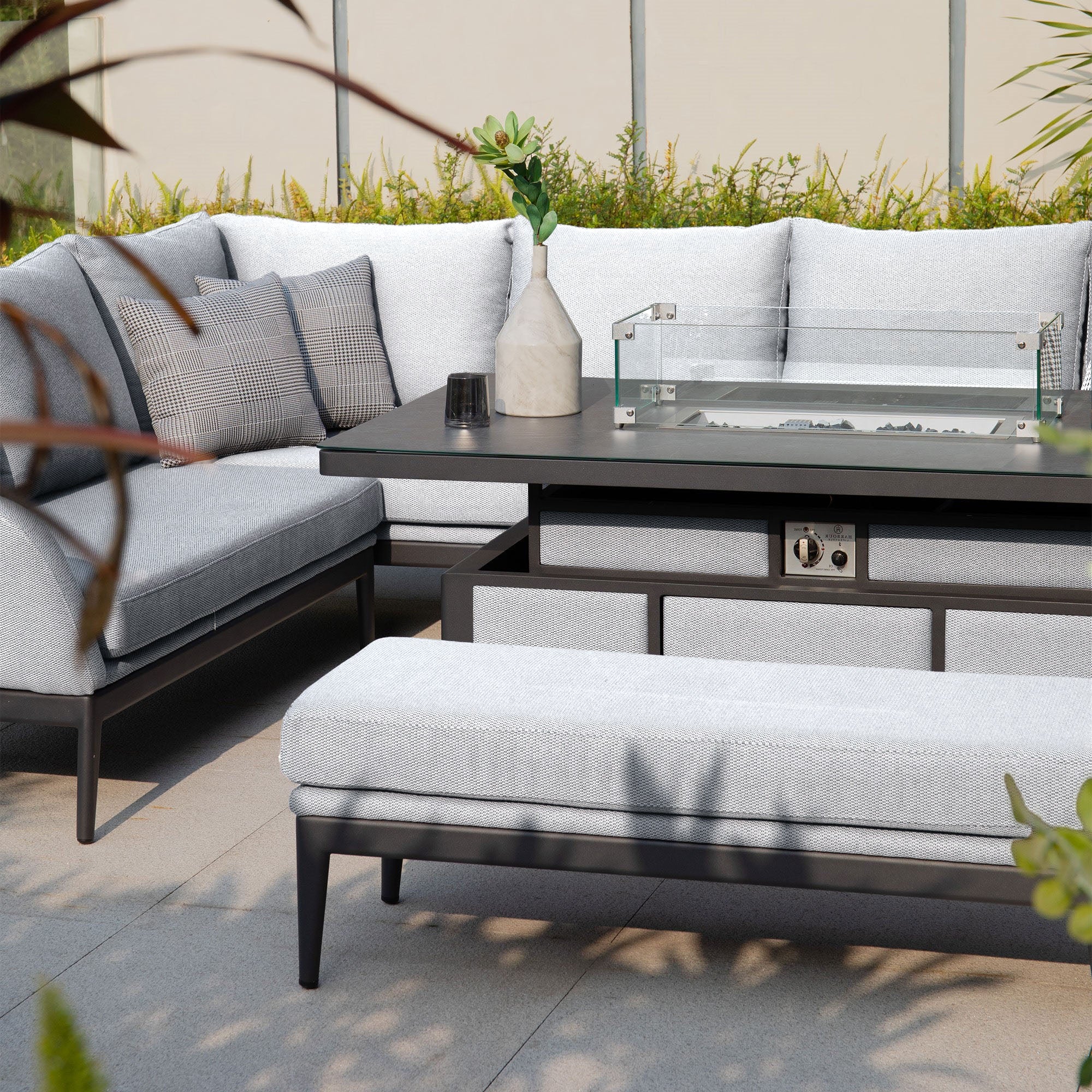 Step into a maintenance-free retreat with the Luna Outdoor Fabric Rectangular Corner Dining Set with Rising Firepit Table in Oyster Grey (Left Hand). This outdoor lounge set features light gray cushions, creating a cozy corner sofa. The set comes complete with a rising firepit table adorned with a glass cover and is complemented by various plants for an inviting ambiance.