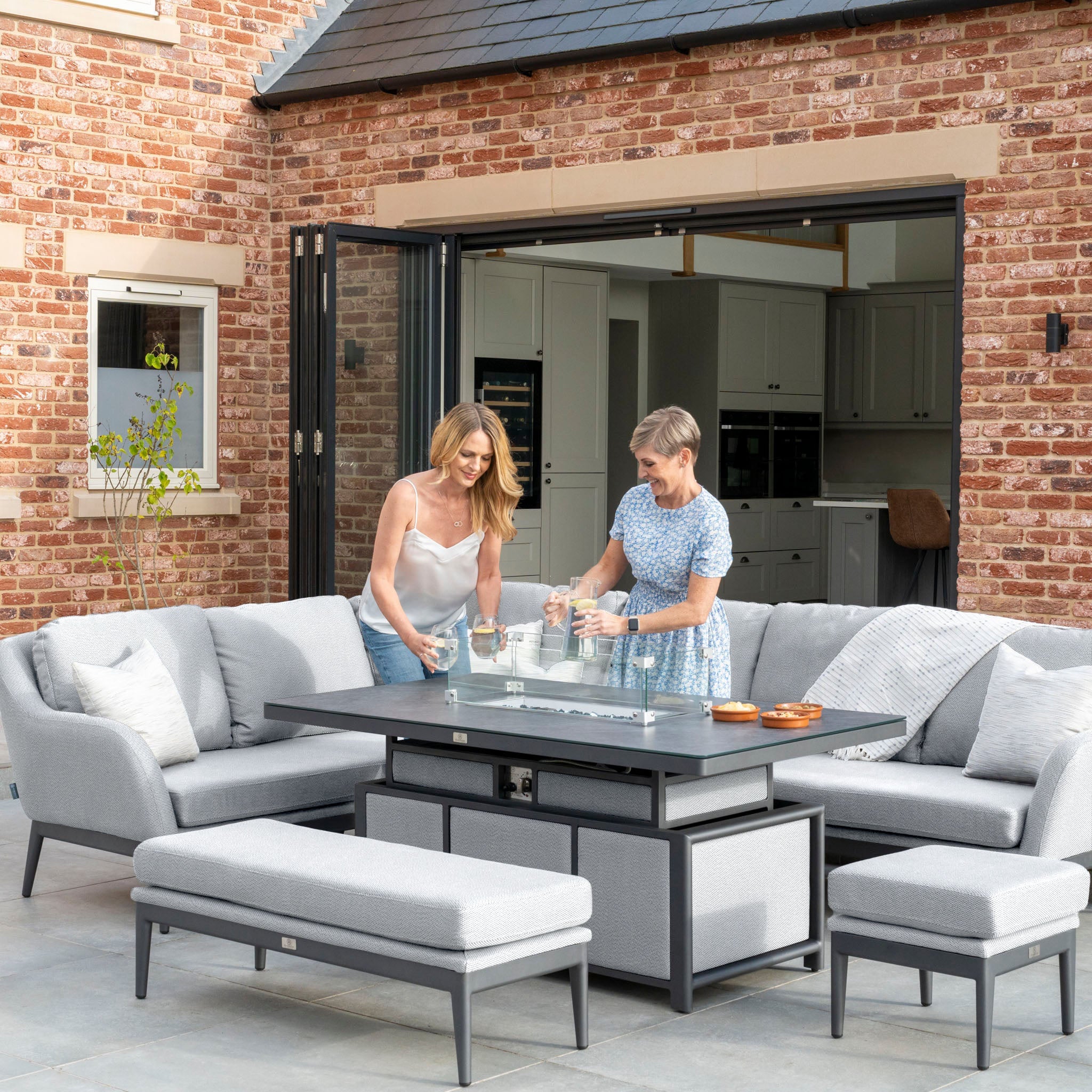 Two women setting up the Luna Outdoor Fabric Rectangular Corner Dining Set with a Rising Firepit Table in Oyster Grey (Left Hand) on a patio, featuring a modern brick house in the background.