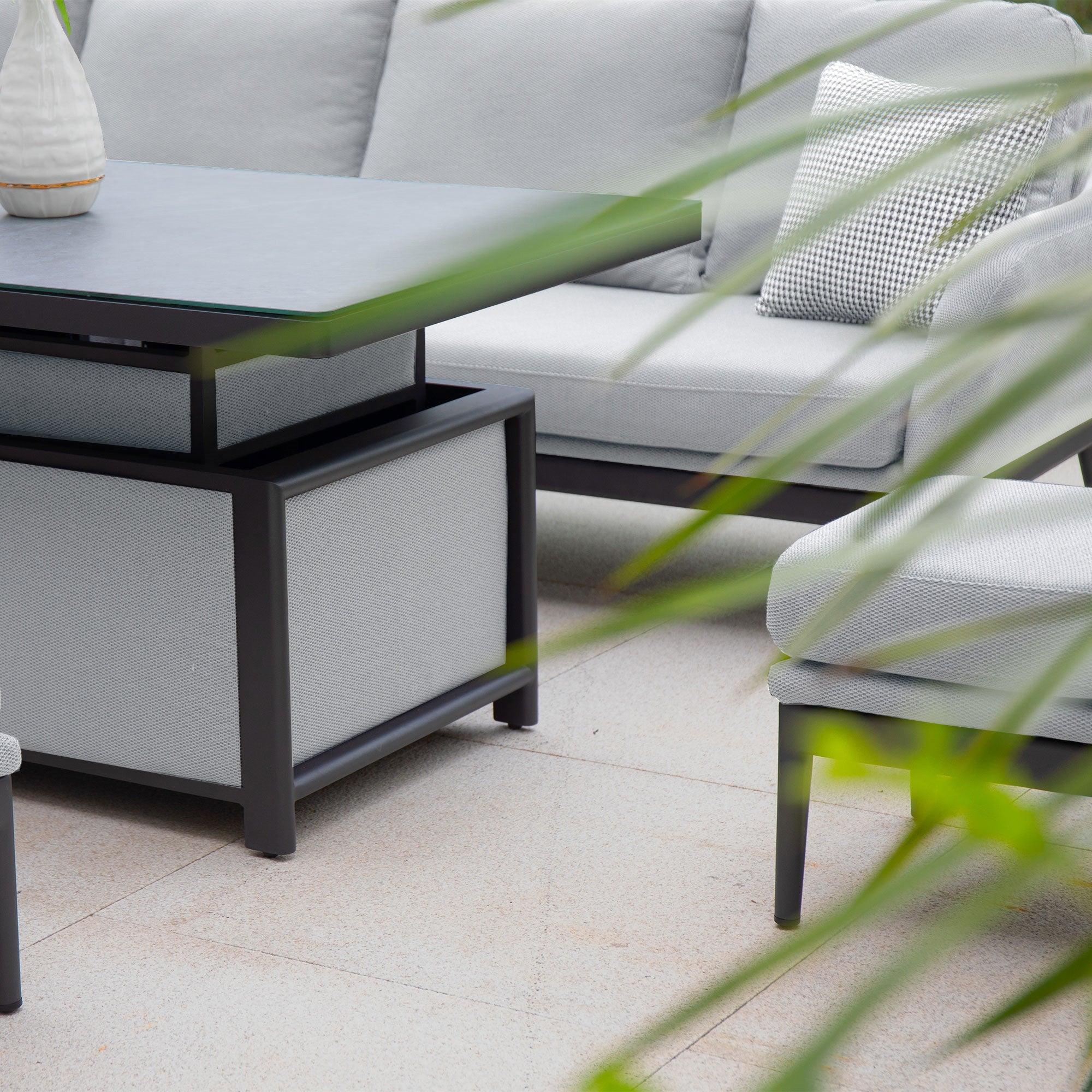 Luna Outdoor Fabric Rectangular Corner Dining Set with Rising Table in Oyster Grey (Left Hand), featuring durable cushions and a rectangular table, partially obscured by green leaves in the foreground. This maintenance-free dining set is perfect for outdoor enjoyment.