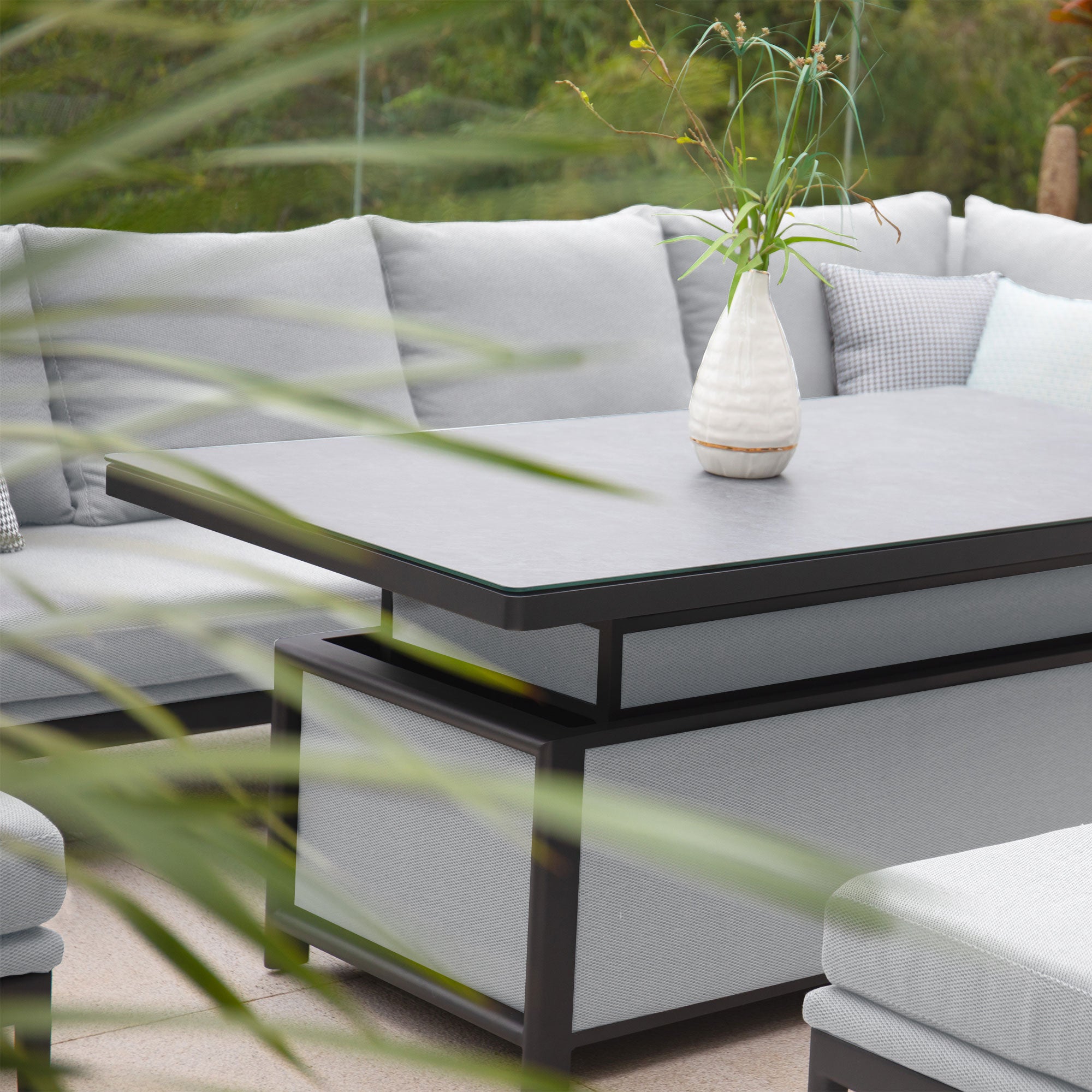 Modern outdoor patio set featuring durable Luna Outdoor Fabric in oyster grey, complete with a rectangular corner dining set in a left-hand configuration and a rising table adorned with a white vase and greenery on top.