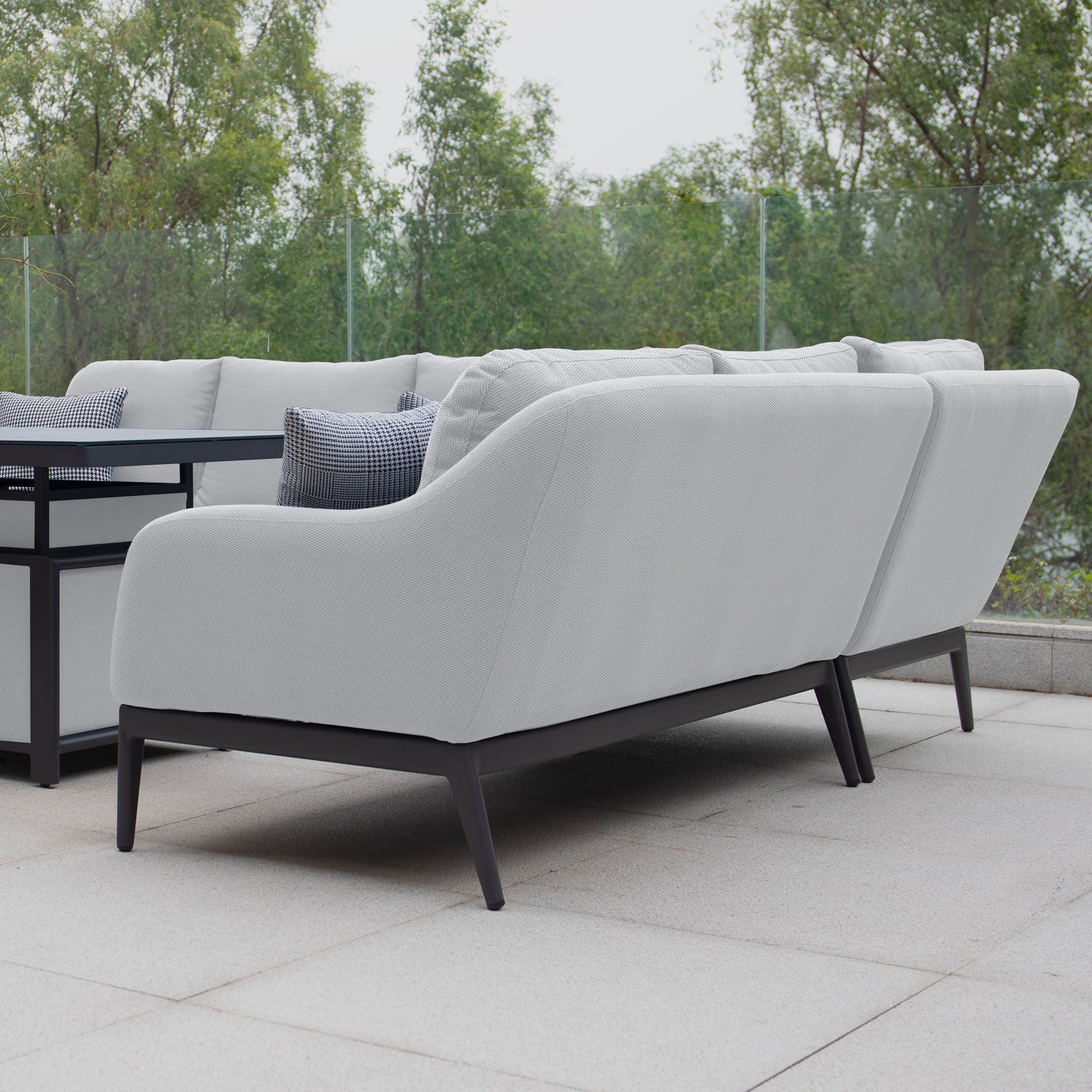 Outdoor seating area with cushioned sofas in durable Luna outdoor fabric (Oyster Grey) and a glass-topped black metal rising firepit table on a tiled patio, surrounded by trees, featuring the Luna Outdoor Fabric Rectangular Corner Dining Set with Rising Firepit Table (Left Hand).