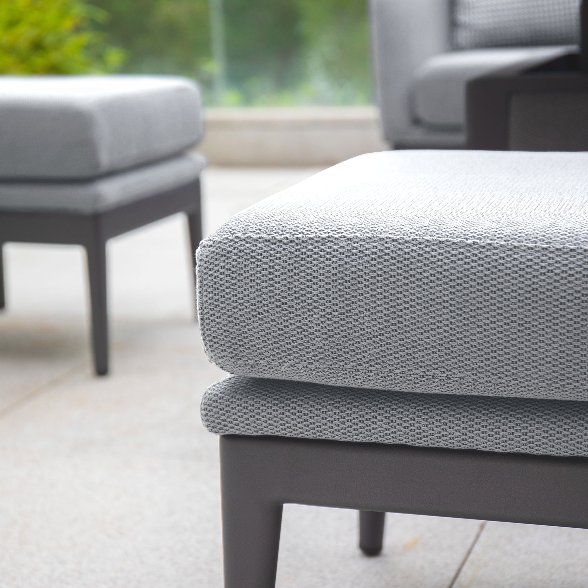 Close-up of a maintenance-free, modern outdoor furniture set, featuring gray Luna cushions on the Luna Outdoor Fabric Rectangular Corner Dining Set with Rising Table in Oyster Grey (Left Hand), positioned on a patio against a blurred green background.