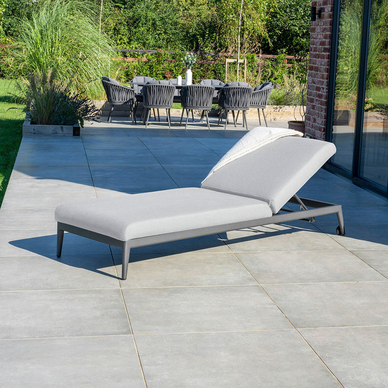 An Oyster Grey Luna Outdoor Fabric Sun Lounger, crafted from all-weather material, rests on a tiled patio next to a brick wall, with a dining table and chairs in the background.