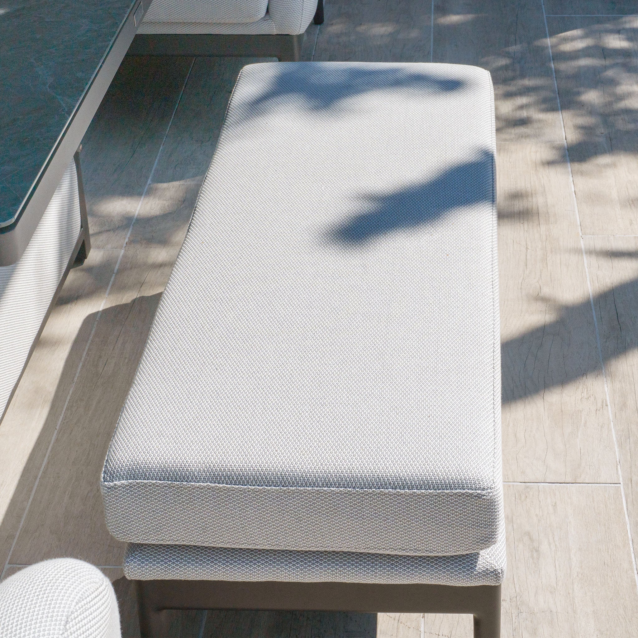 A cushioned ottoman on a sunlit patio casts shadows from nearby trees, perfectly paired with the Luna U-Shape Outdoor Fabric Corner Dining Set with Rising Table in Oyster Grey.