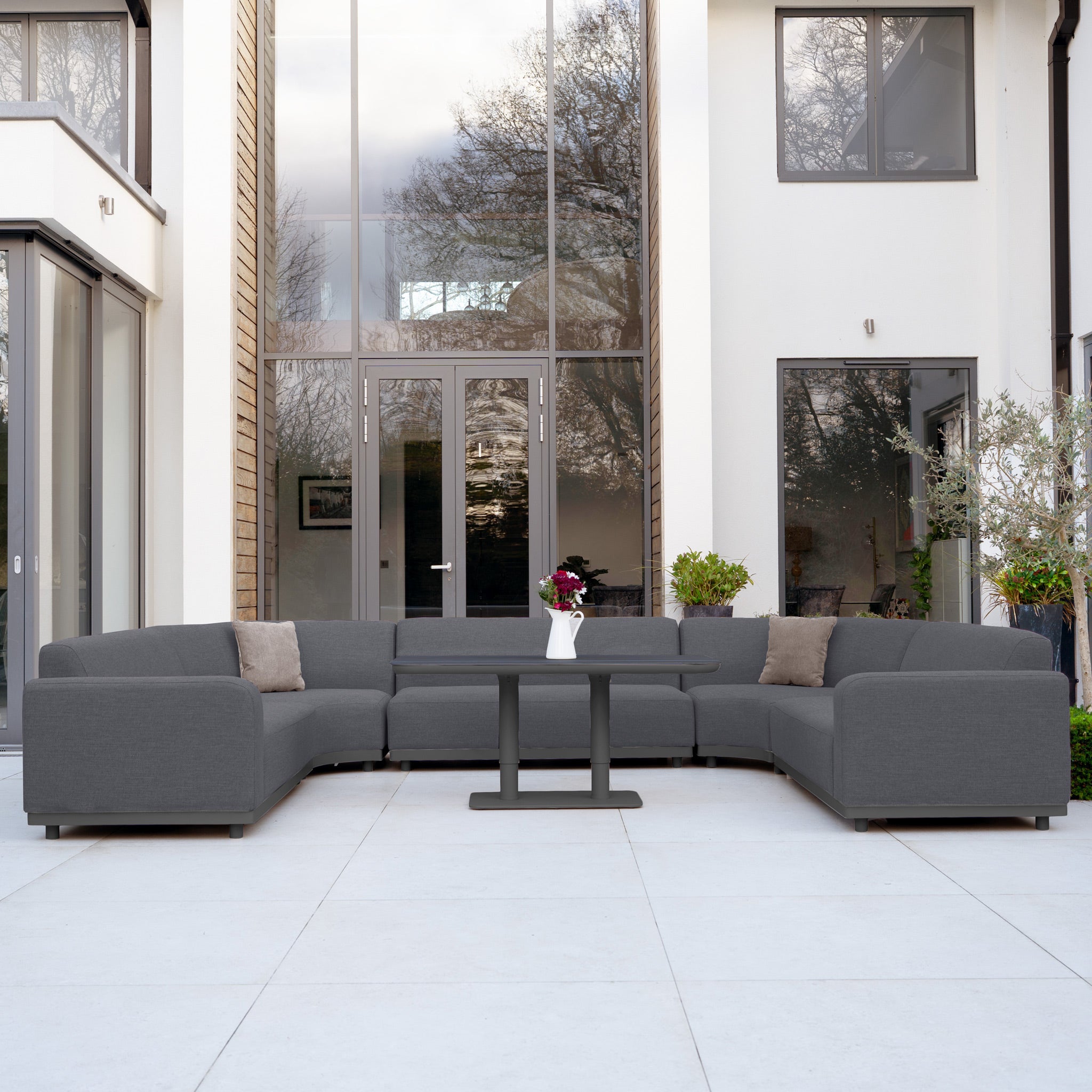 The Luna Outdoor Fabric Curved U-Shape Sofa Set with Altura Rising Table in Grey is stylish all-weather garden furniture, ideal for relaxing on a modern patio in front of large glass doors and windows.