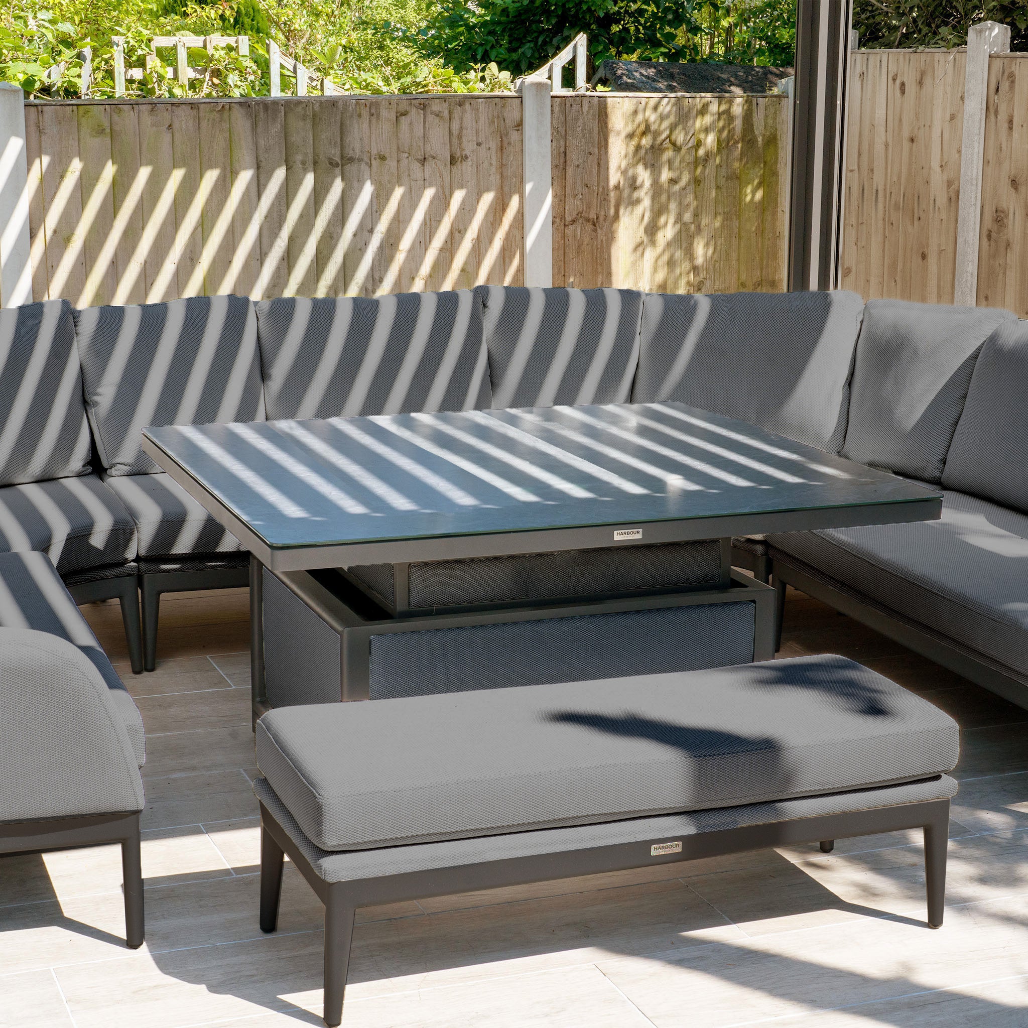 Outdoor patio showcasing the Luna U-Shape Outdoor Fabric Corner Dining with Rising Table in Grey, complete with gray Luna outdoor fabric cushions and a large rising firepit table under sunlit wooden slats.
