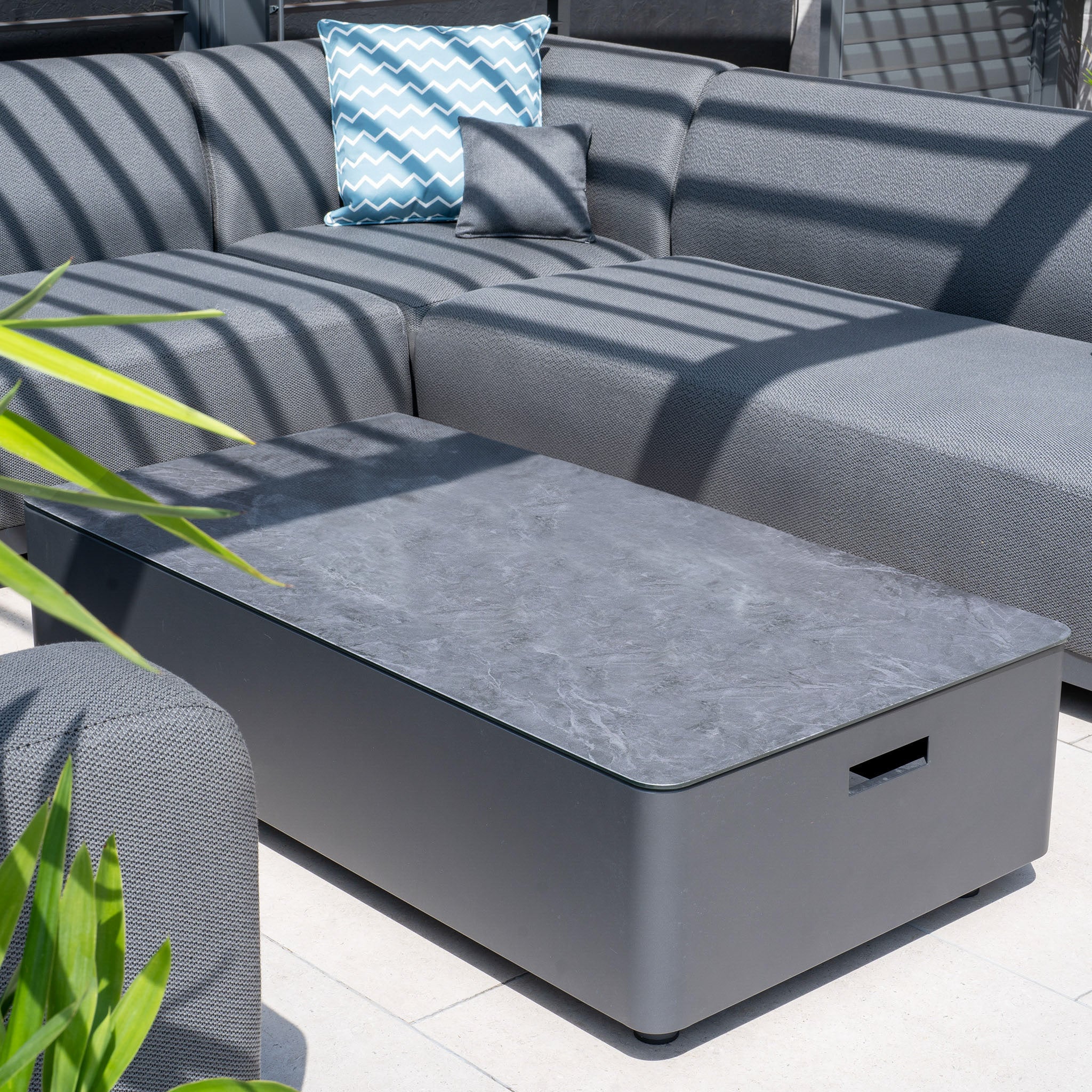 Enjoy the Luna U-Shape Outdoor Fabric Sofa Set with its grey, maintenance-free sectional and patterned cushions, paired perfectly with a modern black coffee table under striped shadows.