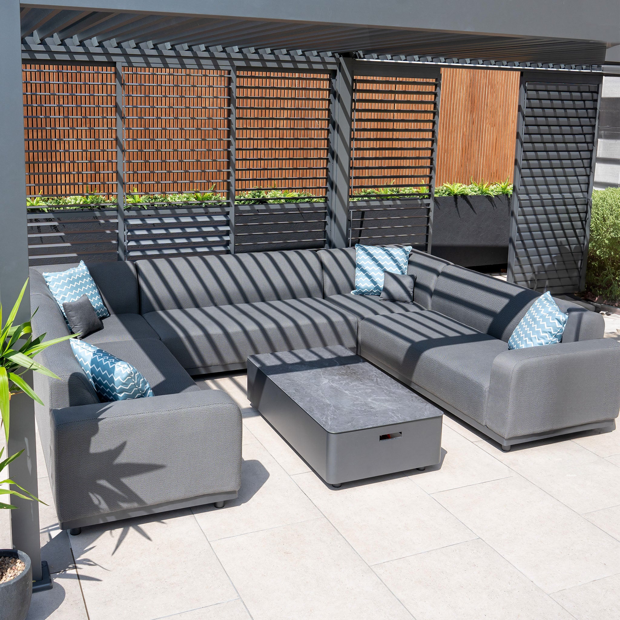 Contemporary outdoor patio featuring the Luna U-Shape Outdoor Fabric Sofa Set in Grey with blue patterned cushions, a matching coffee table, and a slatted wooden screen overhead. This maintenance-free setup guarantees lasting comfort and style.
