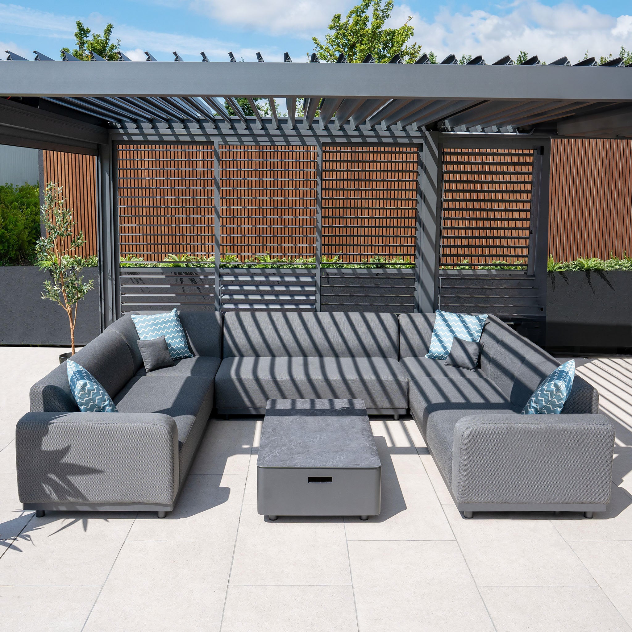 The outdoor seating area includes the Luna U-Shape Outdoor Fabric Sofa Set with Coffee Table in Grey positioned under a pergola, adorned with blue and white pillows. This stylish furniture set features quick-dry foam cushions, ensuring both comfort and elegance regardless of the weather.