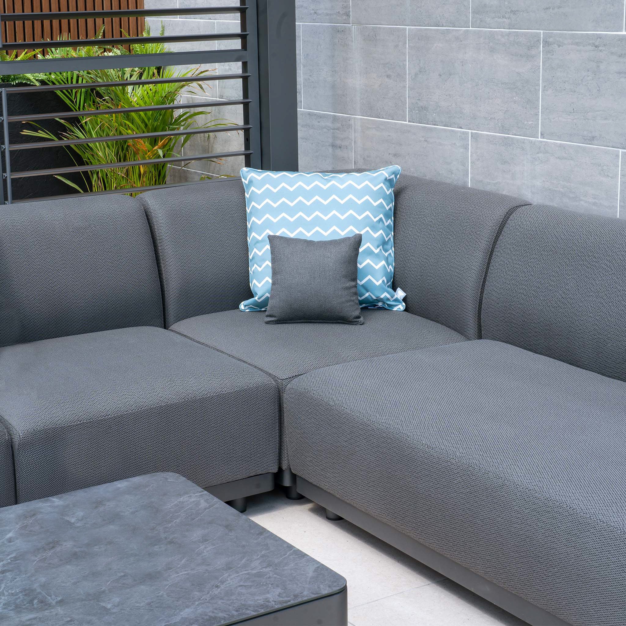 Outdoor patio with the maintenance-free, all-weather proof Luna U-Shape Outdoor Fabric Sofa Set in gray, which includes two pillows (one blue with a white chevron pattern, one gray) and a matching gray coffee table.