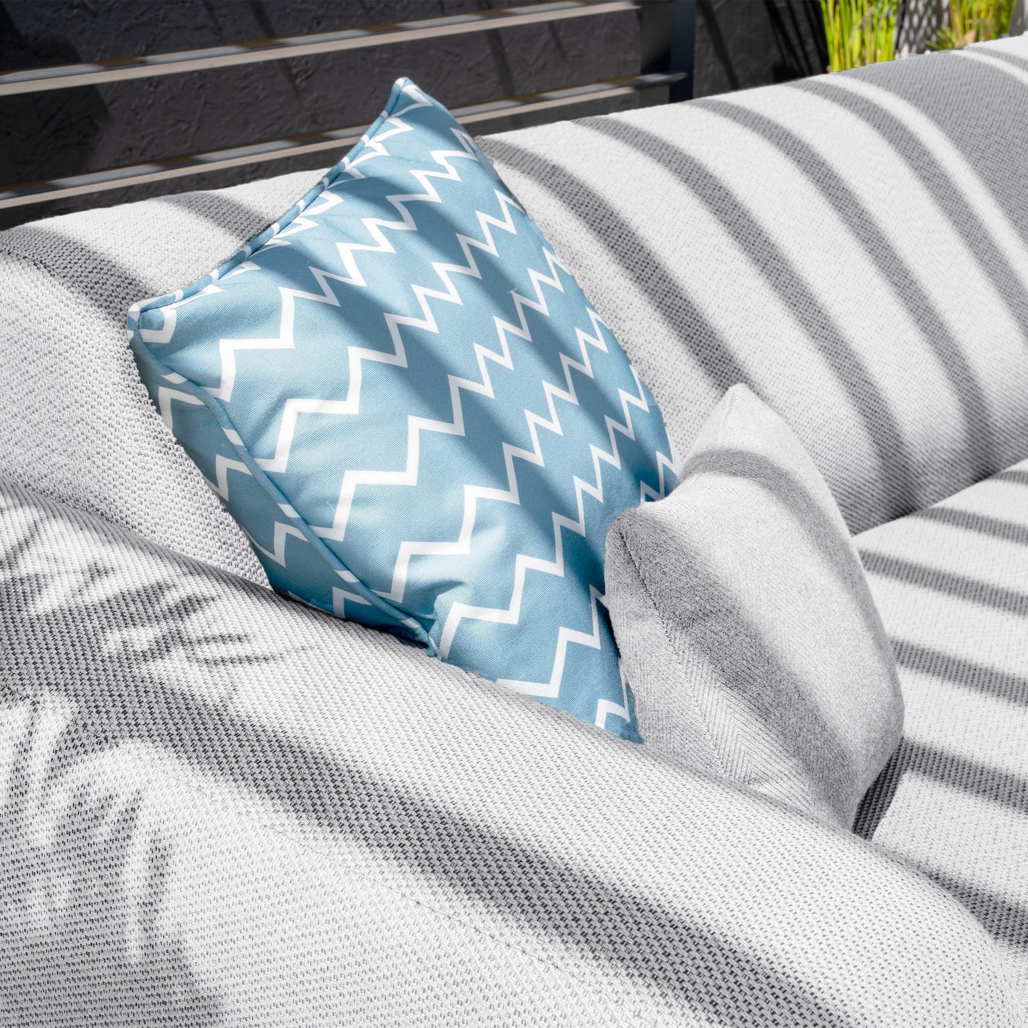 A blue zigzag-patterned pillow made of all-weather material sits on the Luna Outdoor Fabric Large Corner Group Set with Coffee Table in Oyster Grey, casting striped shadows under the sun.