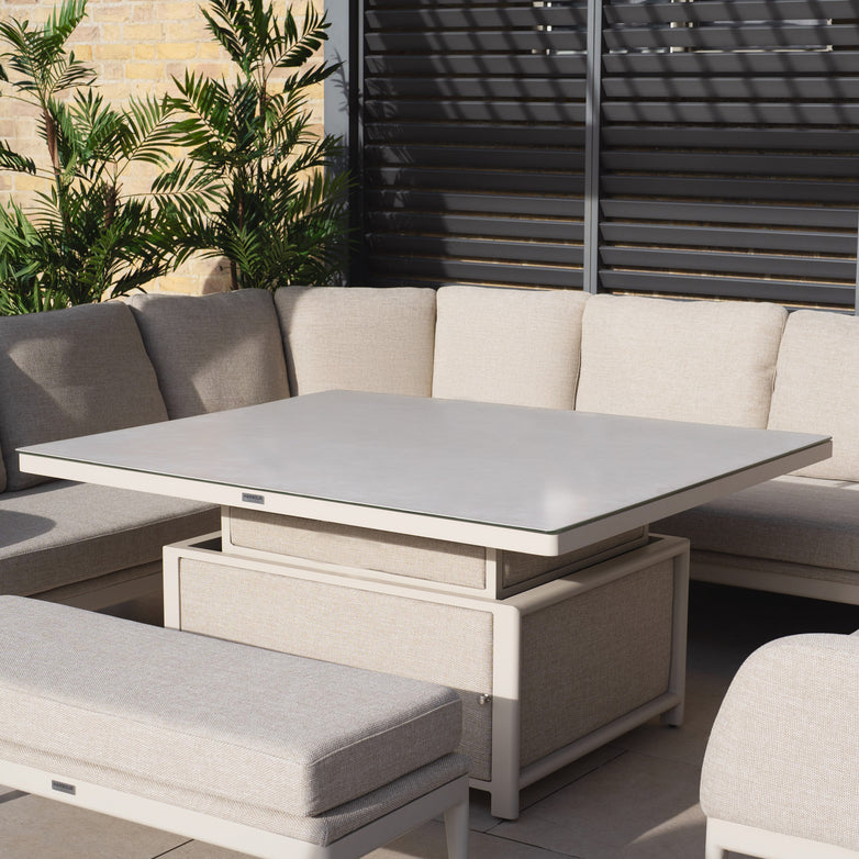 The Luna U-Shape Outdoor Fabric Corner Dining set in Fawn, featuring a rising firepit coffee table, graces a patio beneath slatted panels. Durable plants enhance the setting's charm.