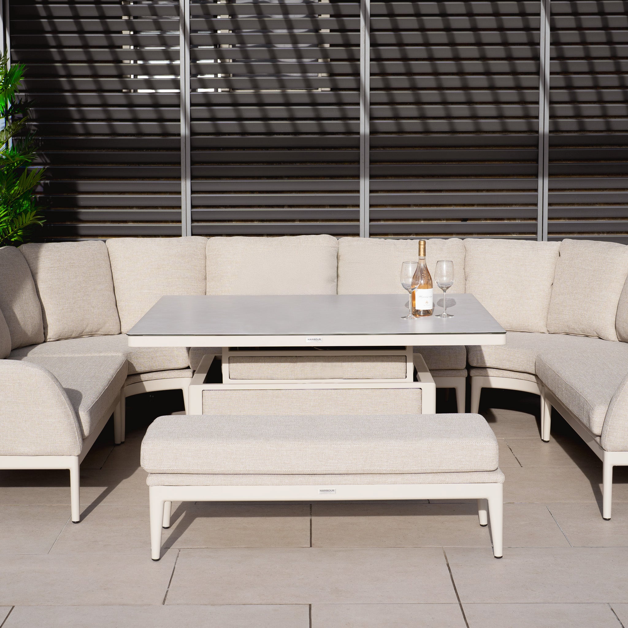 The Luna U-Shape Outdoor Fabric Corner Dining in Fawn includes a stylish sofa, stain-proof table, bench, two wine glasses, and a rising firepit table for an inviting patio setup.