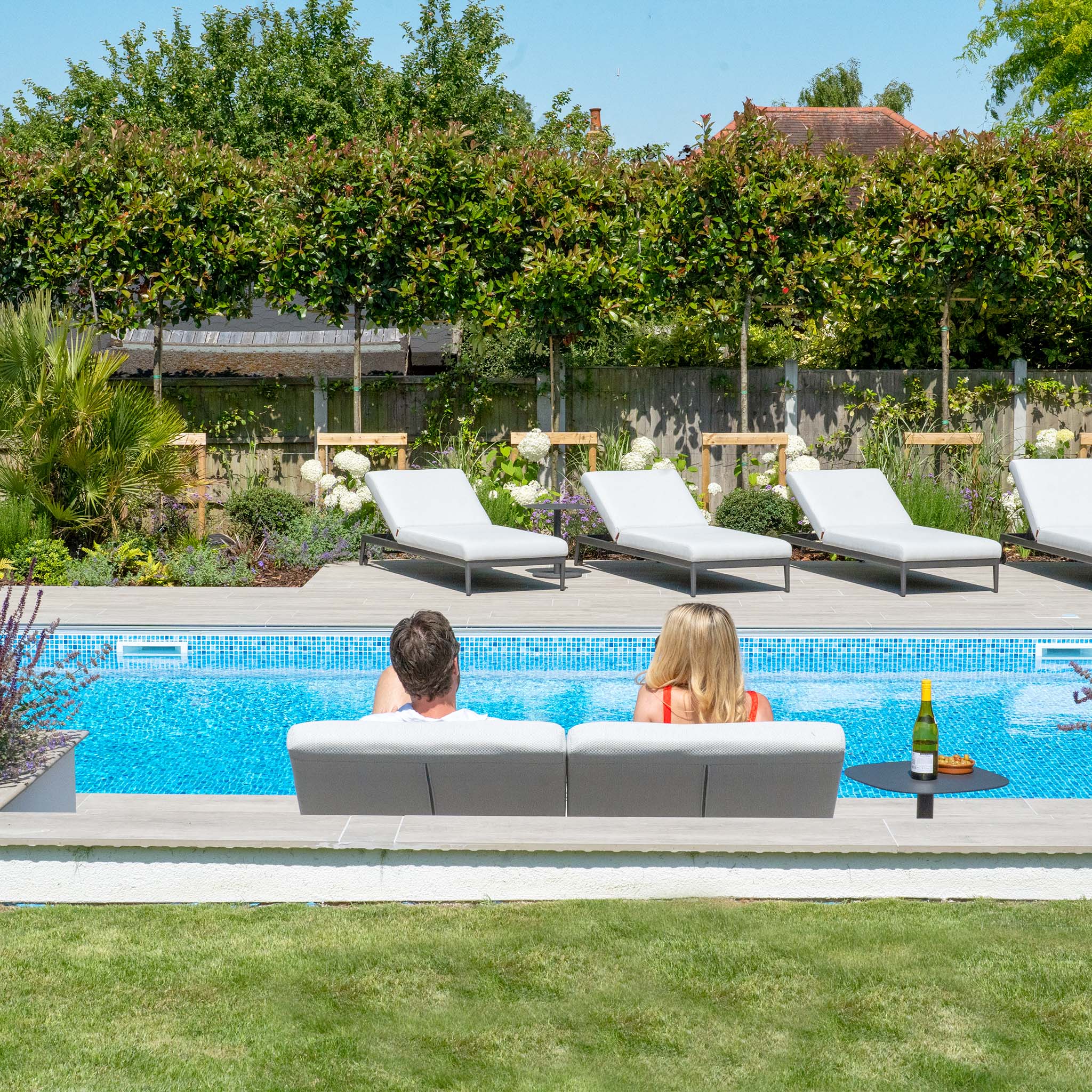 A couple relaxes on a poolside sofa with reflex foam cushions, facing a swimming pool with lounge chairs and trees in the background. This serene setup features the Ali testing vimeo - no video, enhanced by its hydrophobic thin rope weave design.