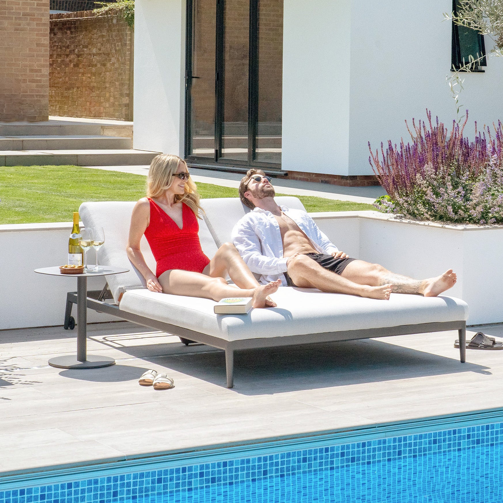 Two people recline on a Luna Outdoor Fabric Double Sun Lounger in Oyster Grey under the warm sun. A bottle of wine, two glasses, and a book rest on a table beside them. The lounger is elegantly upholstered in Luna outdoor fabric, ensuring it remains stain-proof and retains its charm throughout their leisure time.