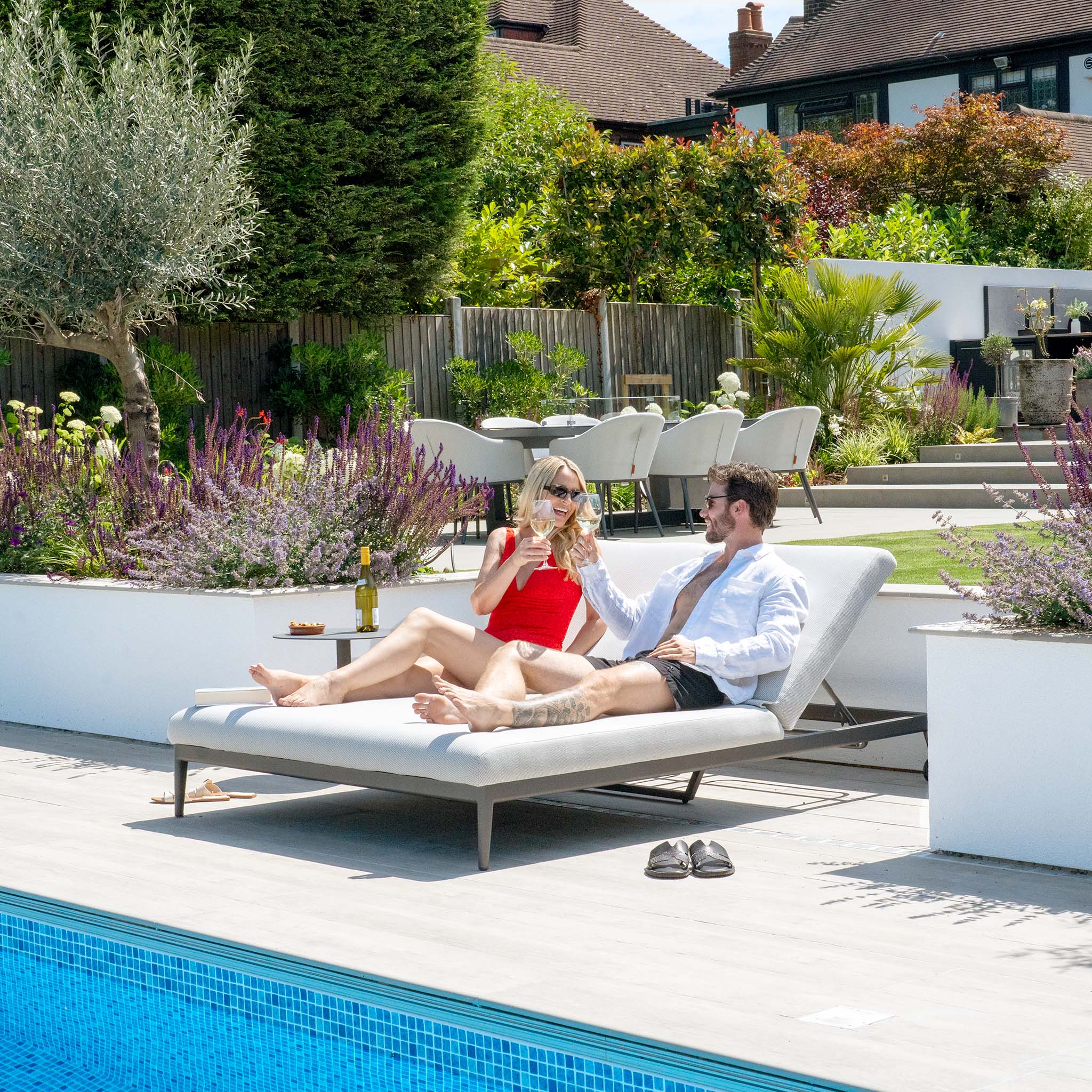 A man and woman relax on the Luna Outdoor Fabric Double Sun Lounger in Oyster Grey, smiling and toasting with drinks in a sunny, landscaped backyard. Their comfort is enhanced by the stain-proof quality of the fabric adorning their lounger.