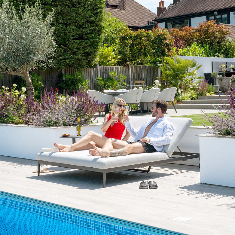 A man and woman relax on the Luna Outdoor Fabric Double Sun Lounger in Oyster Grey, smiling and toasting with drinks in a sunny, landscaped backyard. Their comfort is enhanced by the stain-proof quality of the fabric adorning their lounger.
