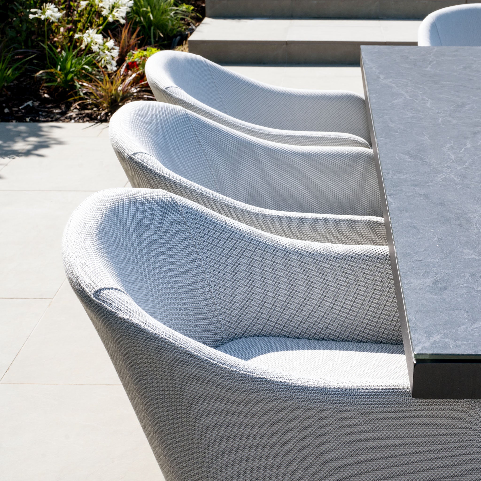 Four cushioned chairs in oyster grey lined up beside a ceramic-topped outdoor table, part of the stylish Luna 8 Seat Outdoor Fabric Rectangular Ceramic Firepit Dining Set, in a sunny patio setting with plants in the background.