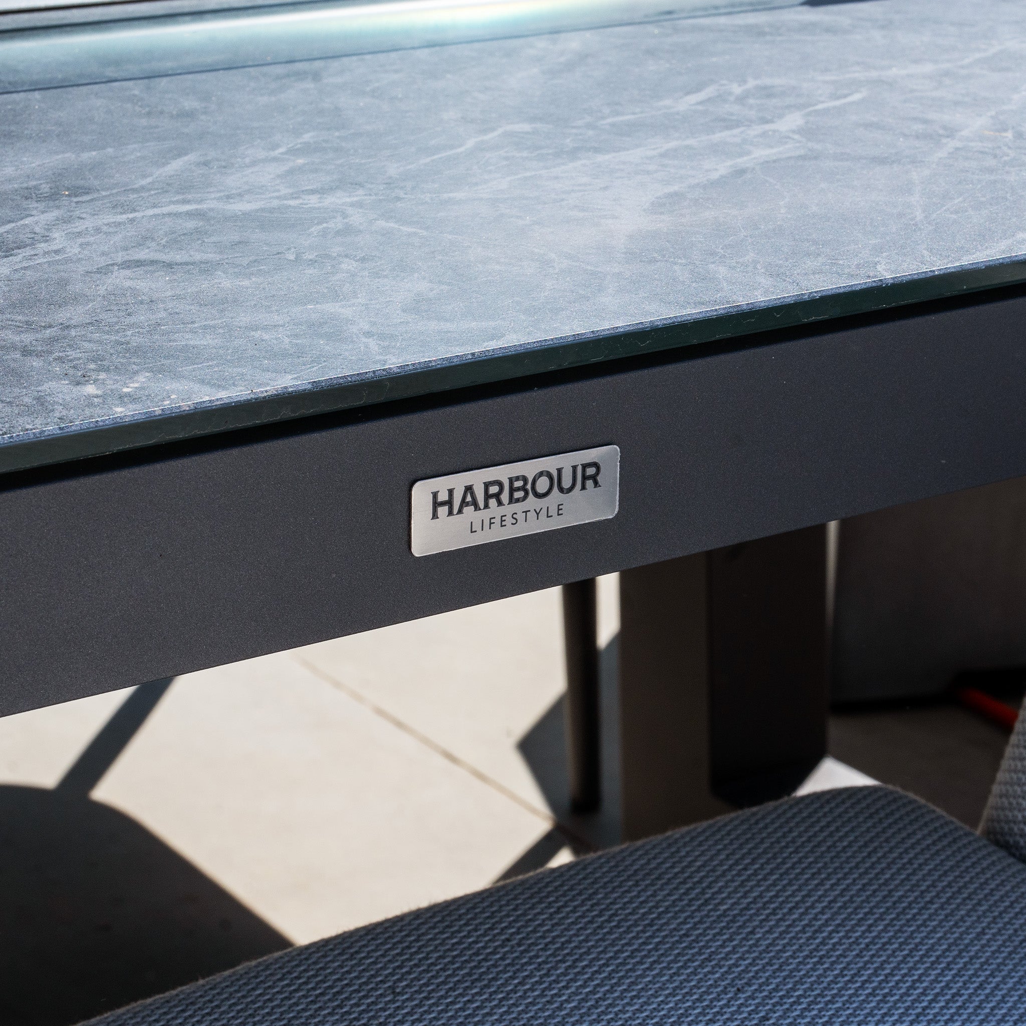 Close-up of a black table with a "Luna 8 Seat Outdoor Fabric Rectangular Ceramic Firepit Dining Set in Oyster Grey" label, set outdoors on a sunny day.