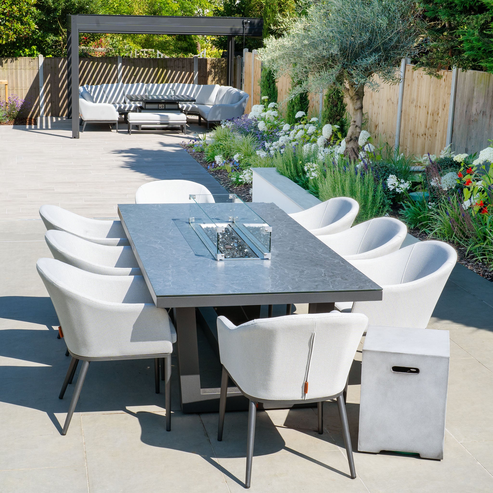 Modern outdoor patio showcasing the Luna 8 Seat Outdoor Fabric Rectangular Ceramic Firepit Dining Set in Oyster Grey, surrounded by a garden with flowers and greenery; seating area in the background.