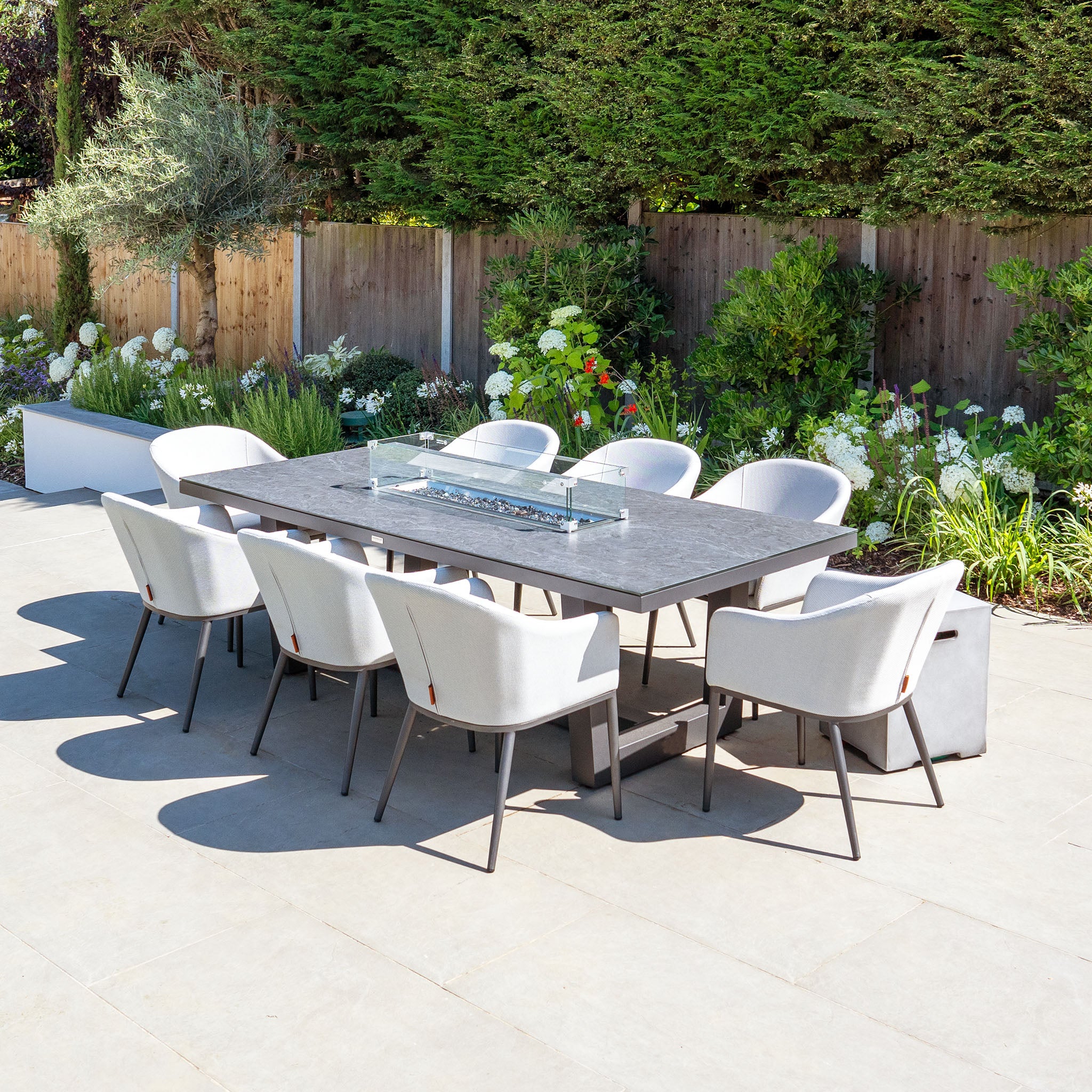 Outdoor dining area showcasing a Harbour Lifestyle Luna 8 Seat Outdoor Fabric Rectangular Ceramic Firepit Dining Set in Oyster Grey, complemented by a garden with lush greenery in the background.