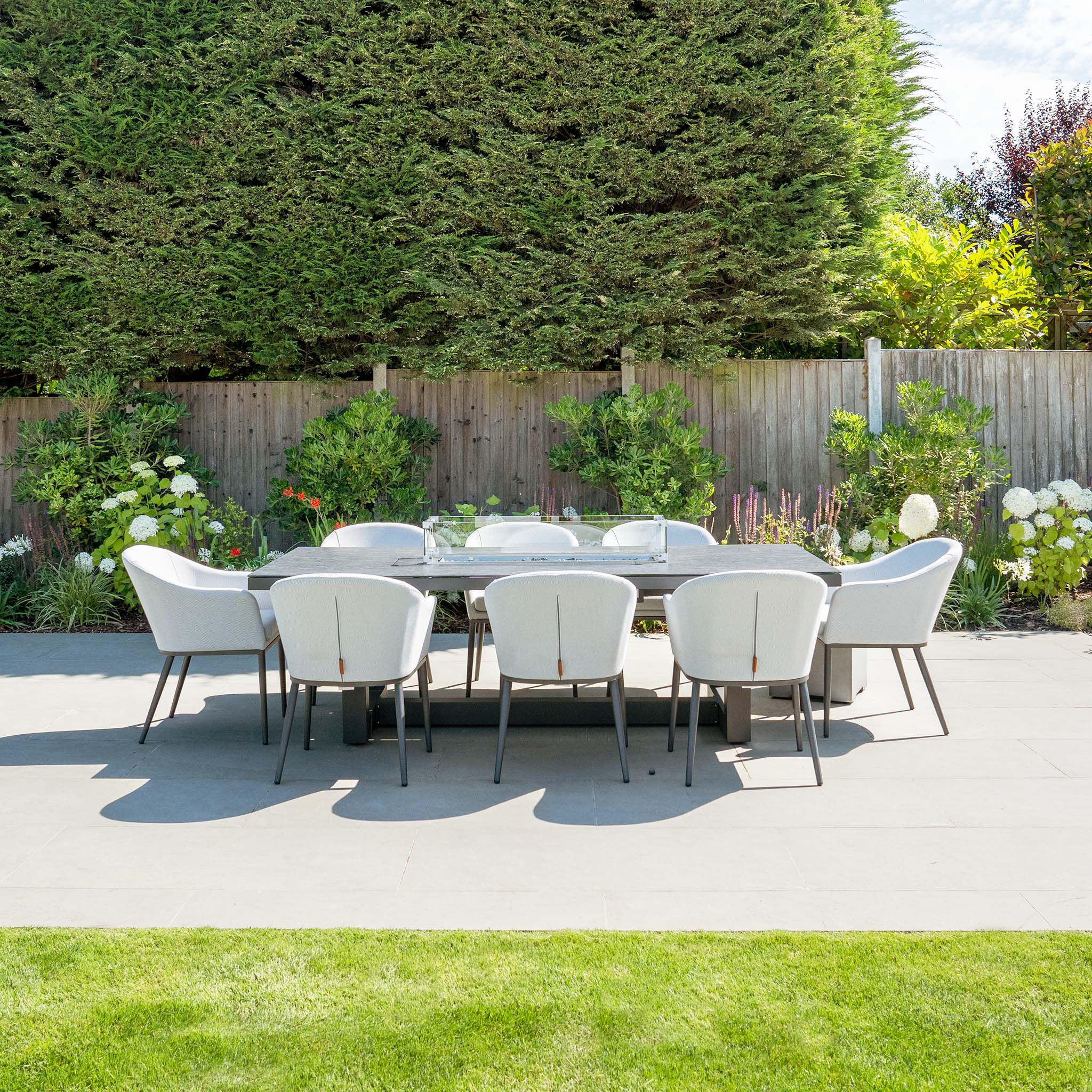 Outdoor patio featuring the Luna 8 Seat Outdoor Fabric Rectangular Ceramic Firepit Dining Set in Oyster Grey, with a large table surrounded by eight white chairs, backed by a wooden fence and greenery.