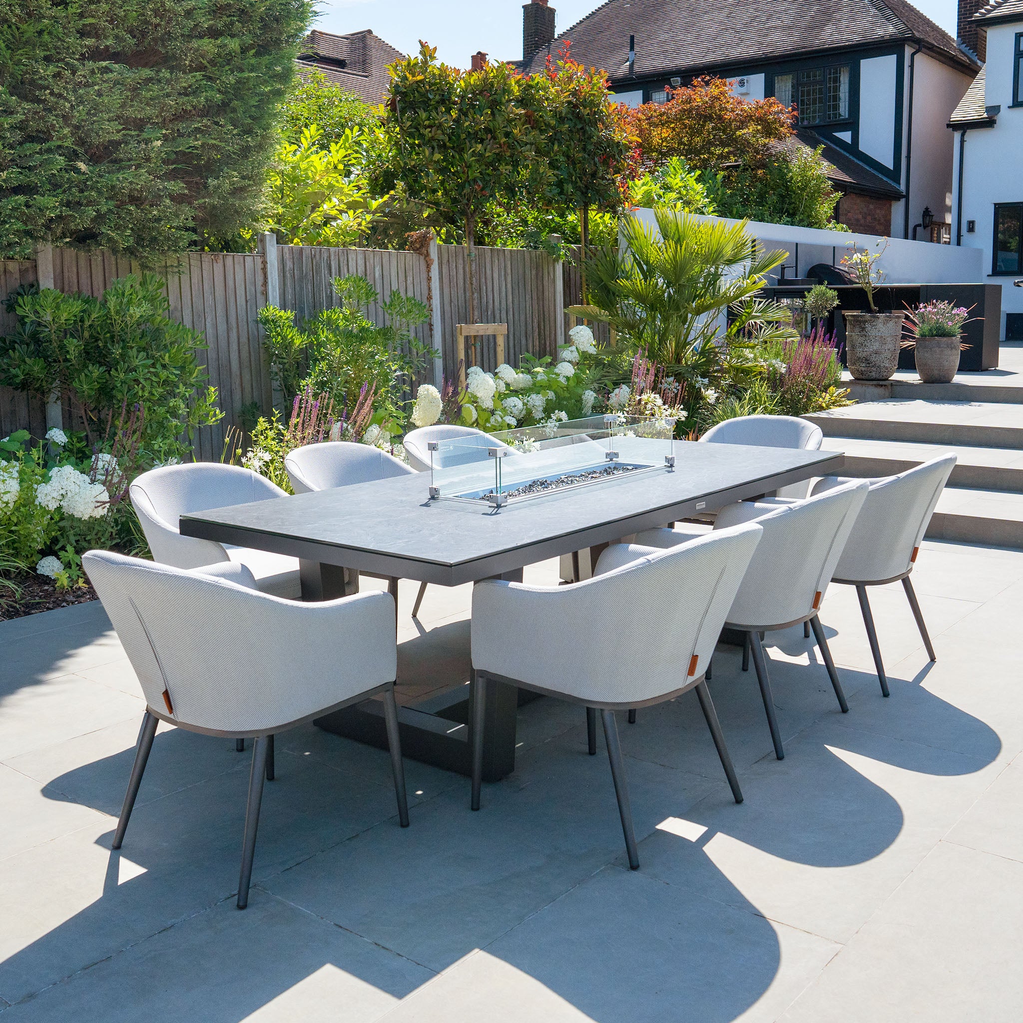 Outdoor dining area with a modern rectangular table firepit set, featuring the Luna 8 Seat Outdoor Fabric Rectangular Ceramic Firepit Dining Set in Oyster Grey by Harbour Lifestyle, surrounded by greenery and a wooden fence.