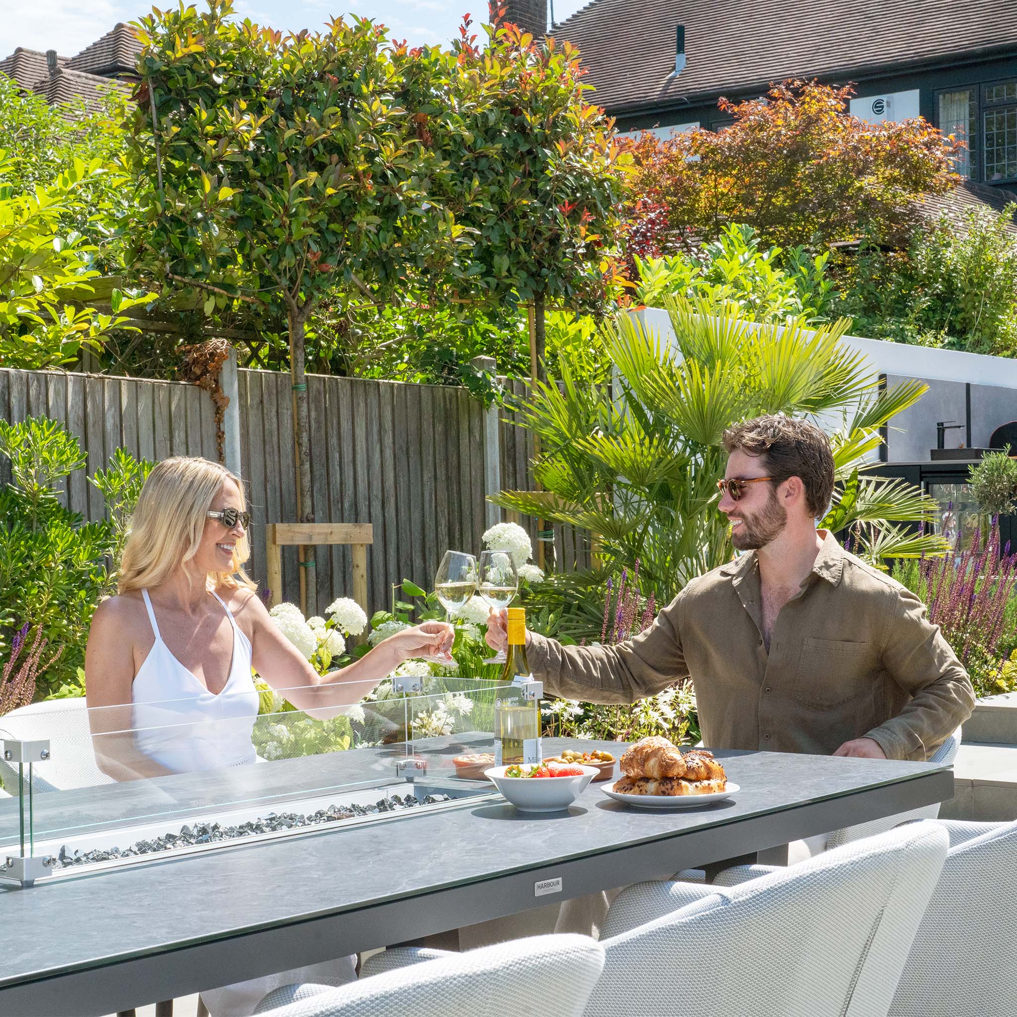 A man and a woman smile while clinking wine glasses at an outdoor patio table with food, surrounded by greenery and seated on the comfortable Ali testing vimeo - no video set.