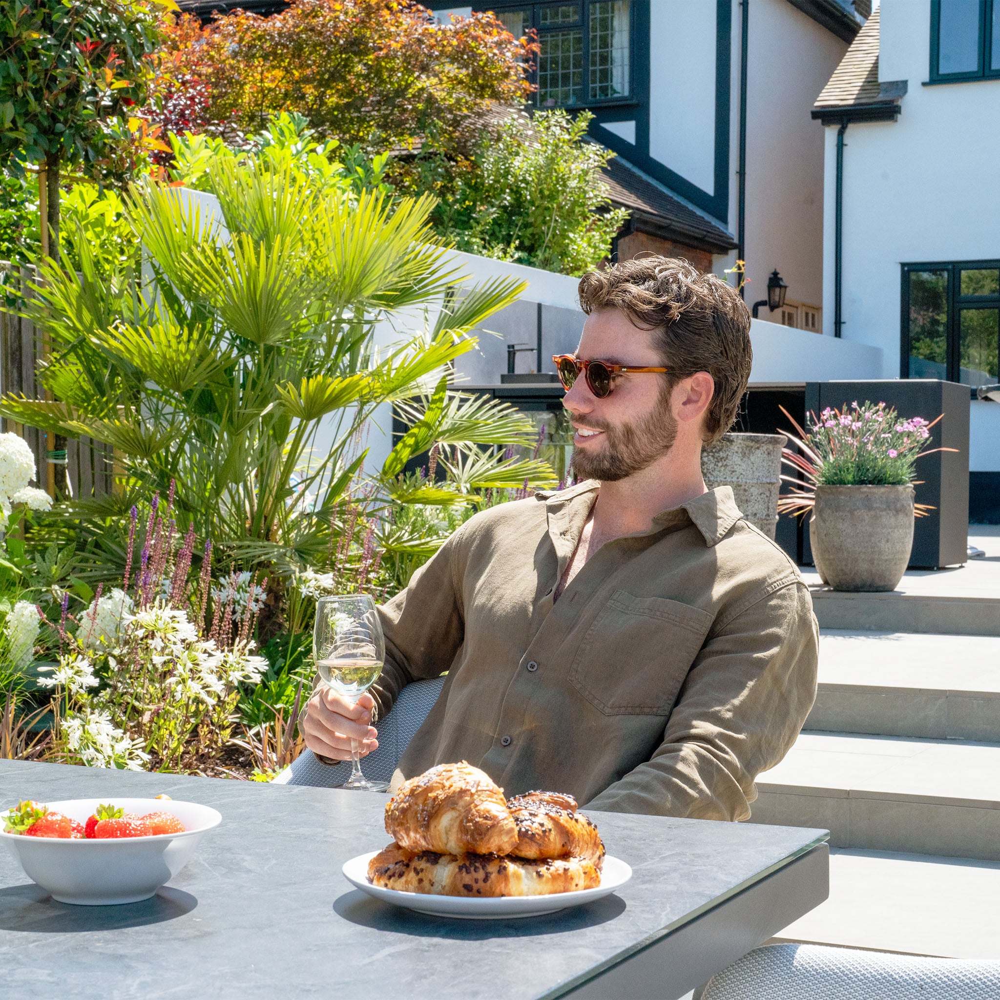 A man wearing sunglasses relaxes comfortably at an outdoor table, enjoying a glass of wine alongside pastries and a bowl of strawberries, seated on the Luna 8 Seat Outdoor Fabric Rectangular Ceramic Firepit Dining Set in Oyster Grey from Harbour Lifestyle.
