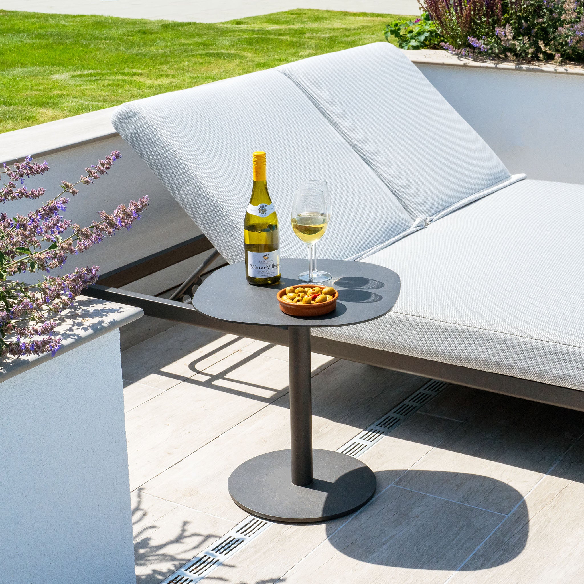 A sunny outdoor lounge area features a Luna Outdoor Fabric Sun Lounger in Oyster Grey from Harbour Lifestyle, accompanied by a bottle of white wine, a glass of wine, and a bowl of olives on a small side table. Made from all-weather materials and Luna outdoor fabric, the lounger promises both comfort and durability.