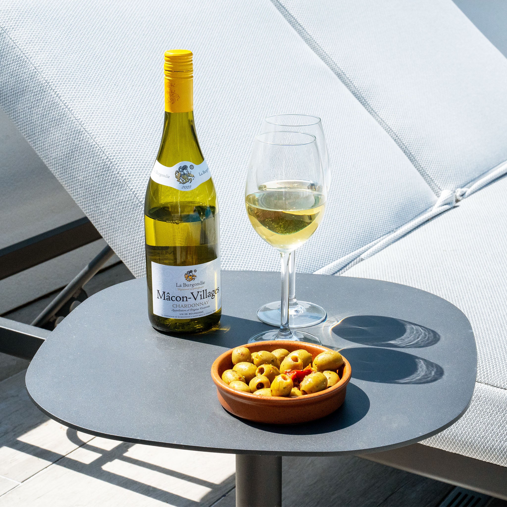 A bottle of white wine, a glass of wine, and a bowl of olives on a table next to the Luna Outdoor Fabric Sun Lounger in Oyster Grey, which boasts all-weather material for ultimate relaxation.