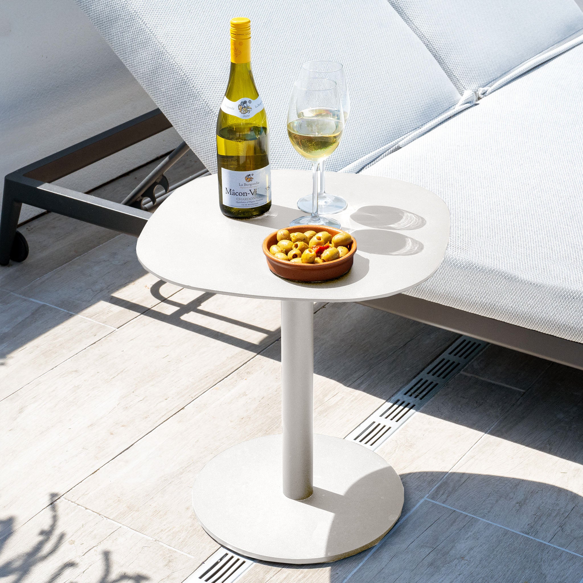 A Luna Side Table in Latte (45cm x 45cm) stands beside a lounge chair, displaying a wine bottle, glass, and bowl of olives—an inviting tableau enhanced by the table’s durable, maintenance-free outdoor design.