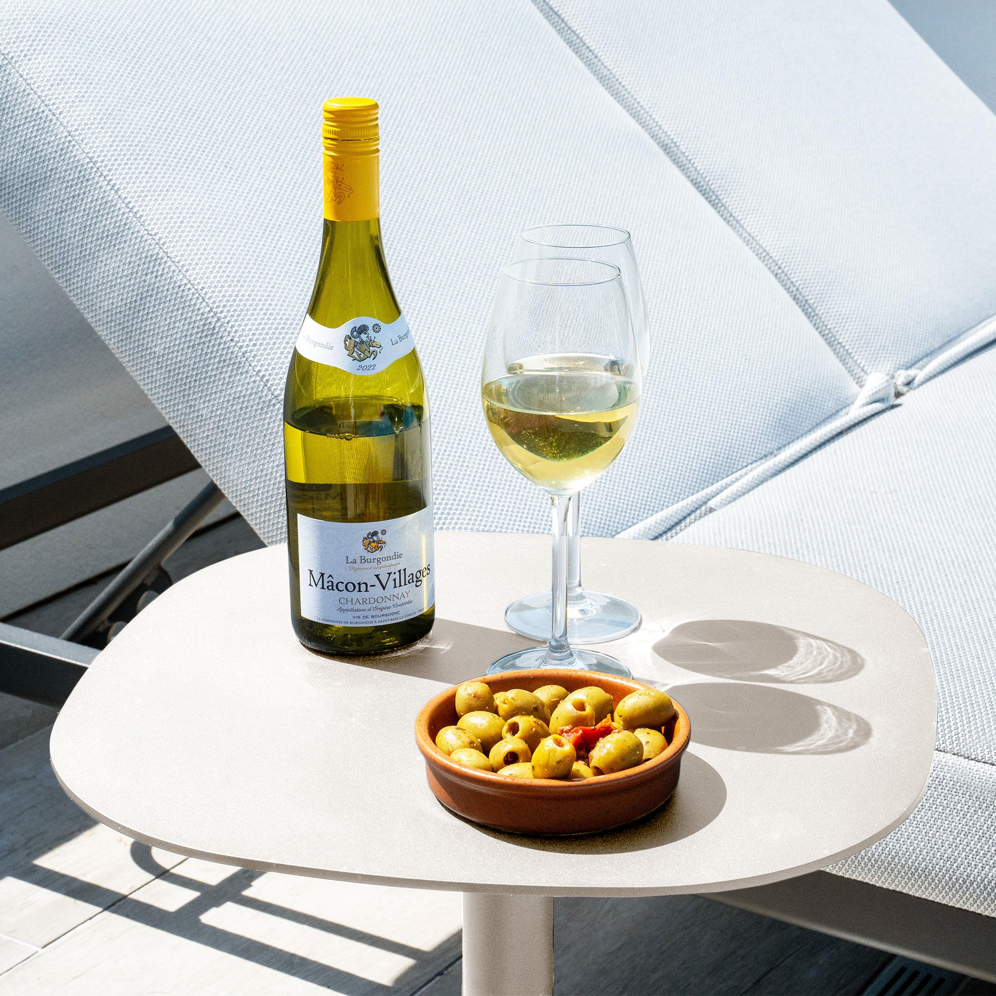 A bottle of white wine, a glass, and a bowl of green olives rest on the Luna Side Table in Latte (45cm x 45cm) outdoors beside weatherproof furniture, all bathed in sunlight by the lounge chair.