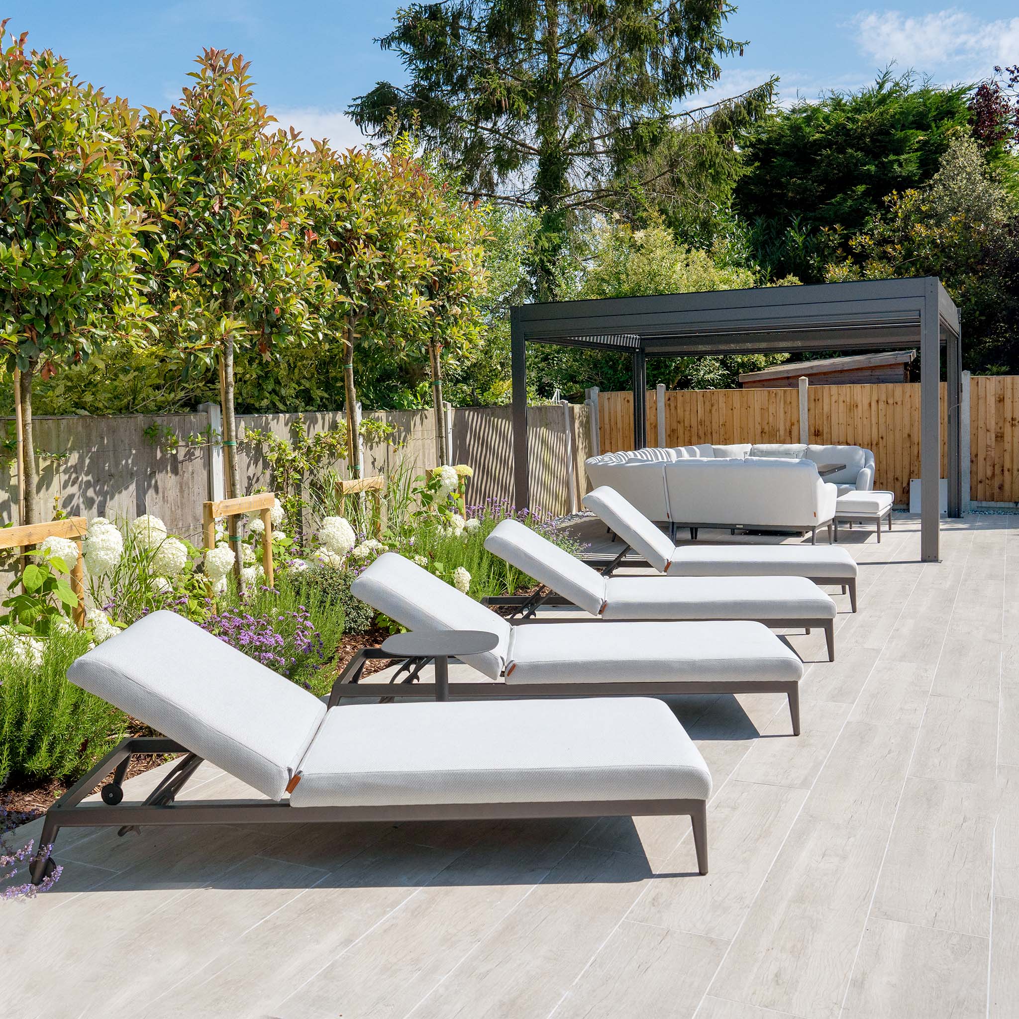 Outdoor patio with multiple Harbour Lifestyle Luna Outdoor Fabric Sun Loungers in Oyster Grey, featuring white cushioned seats in all-weather material and a shaded seating area, surrounded by greenery.