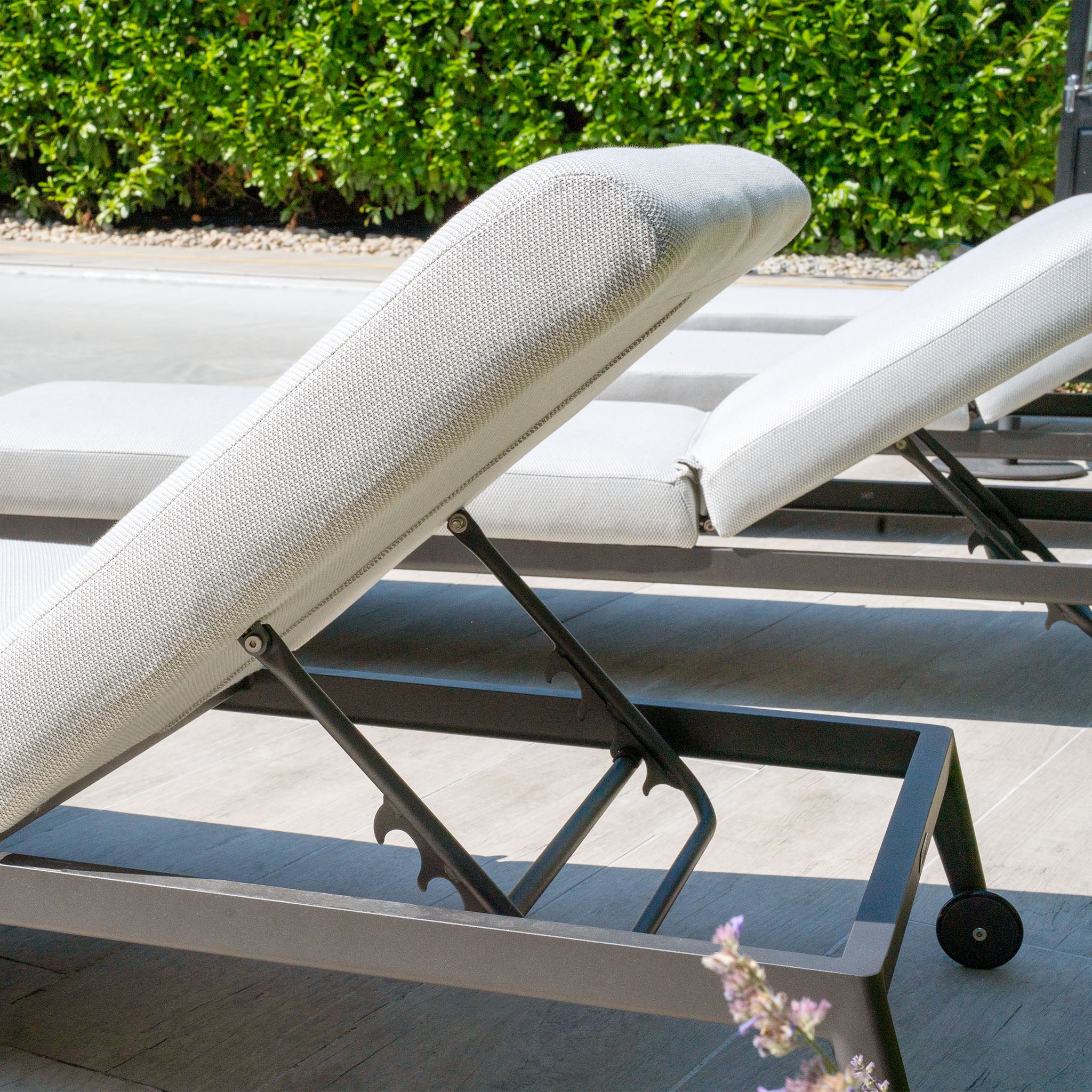The Luna Outdoor Fabric Sun Lounger in Oyster Grey, featuring adjustable backs and crafted from all-weather material, is set on a patio near green hedges.