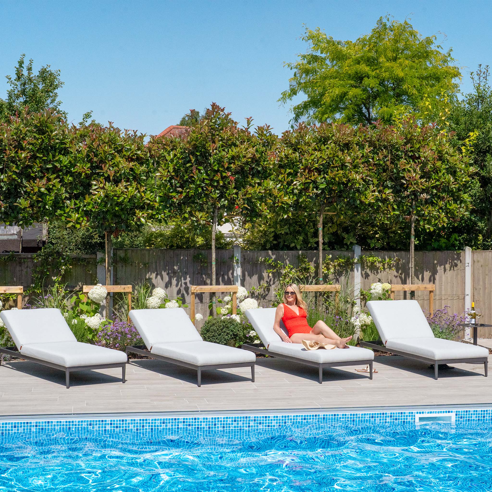 A person relaxes on a Luna Outdoor Fabric Sun Lounger in Oyster Grey beside a pool, surrounded by garden greenery on a sunny day, enjoying the comfort of the all-weather material.