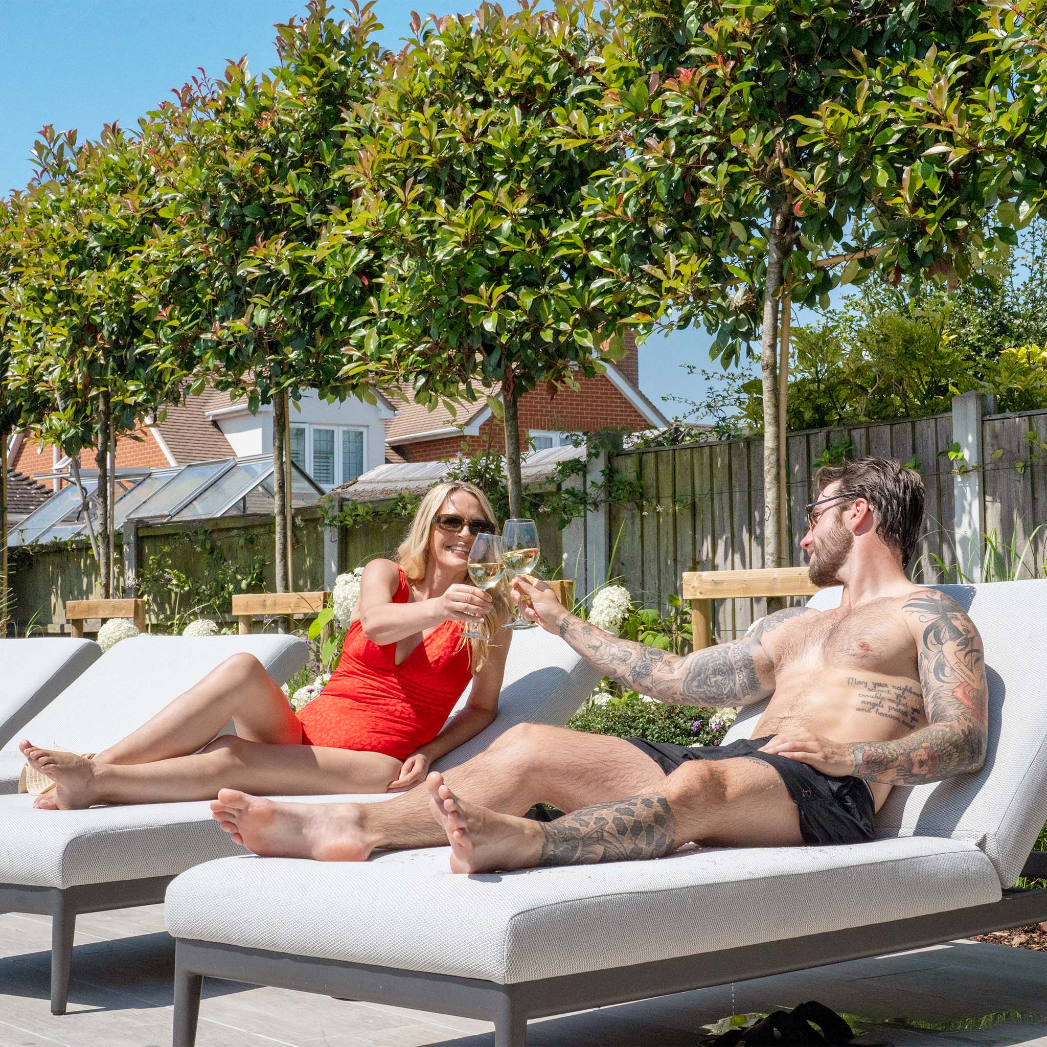 A woman in a red swimsuit and a man with tattoos clink glasses while lounging on Luna Outdoor Fabric Sun Loungers in Oyster Grey outdoors.