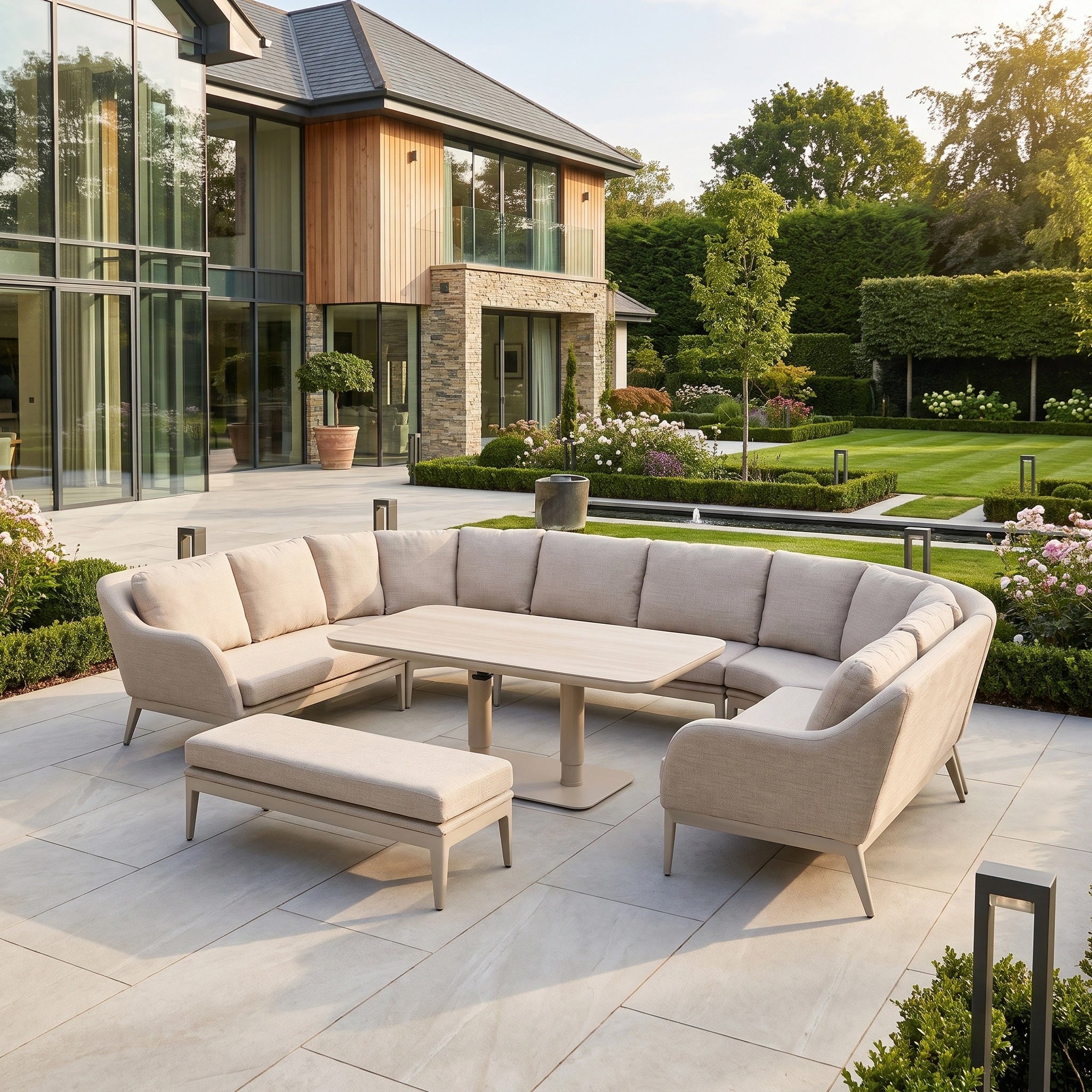 The Luna U-Shape Outdoor Fabric Corner Dining Set with Altura Rising Table in Fawn sits elegantly on a stone patio, backed by a modern house and landscaped garden—ideal for anyone seeking stylish, contemporary outdoor furniture.