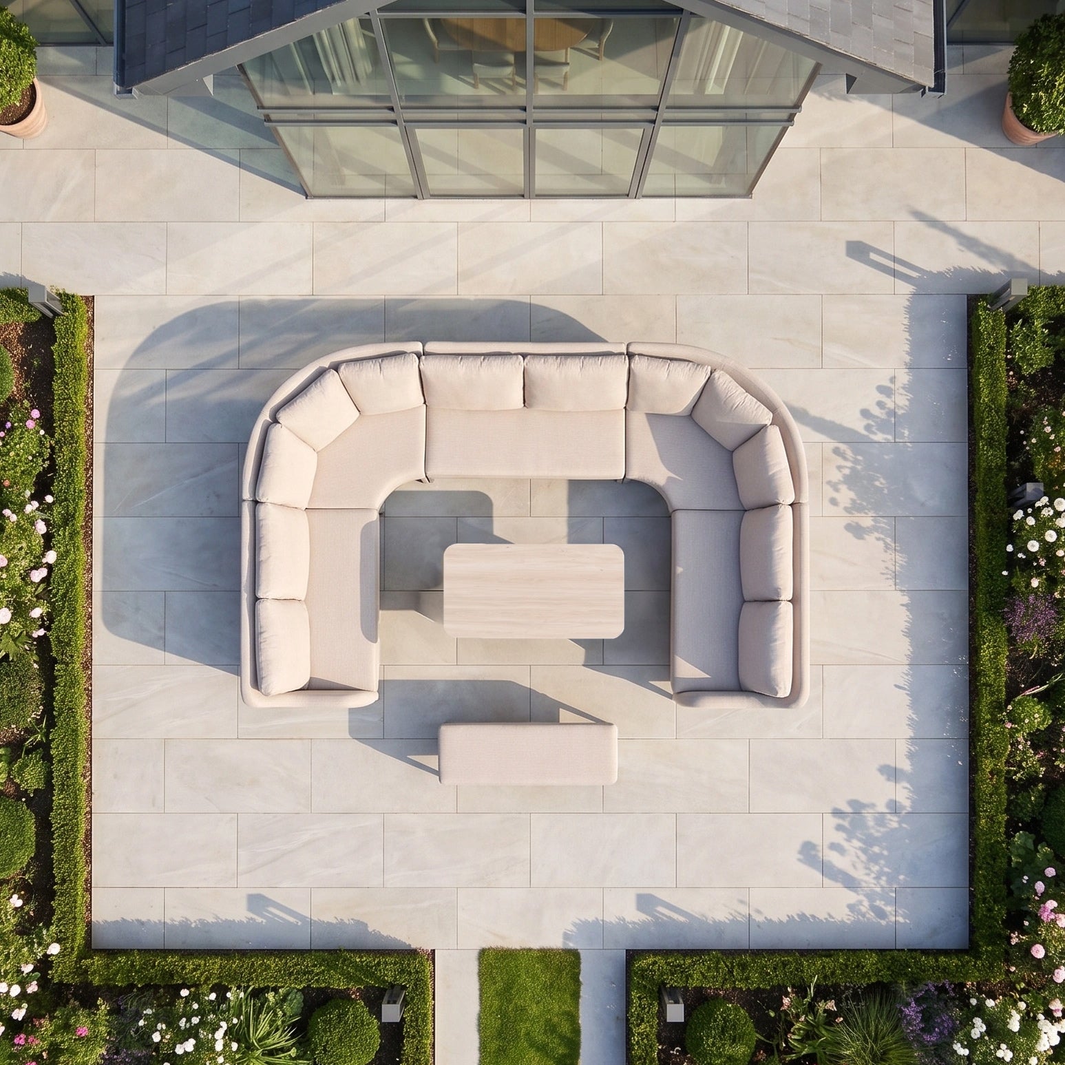 The Luna U-Shape Outdoor Fabric Corner Dining Set with Altura Rising Table in Fawn features a curved beige sofa paired with tables on a tiled patio, surrounded by lush green garden beds, creating a contemporary and inviting outdoor furniture arrangement.