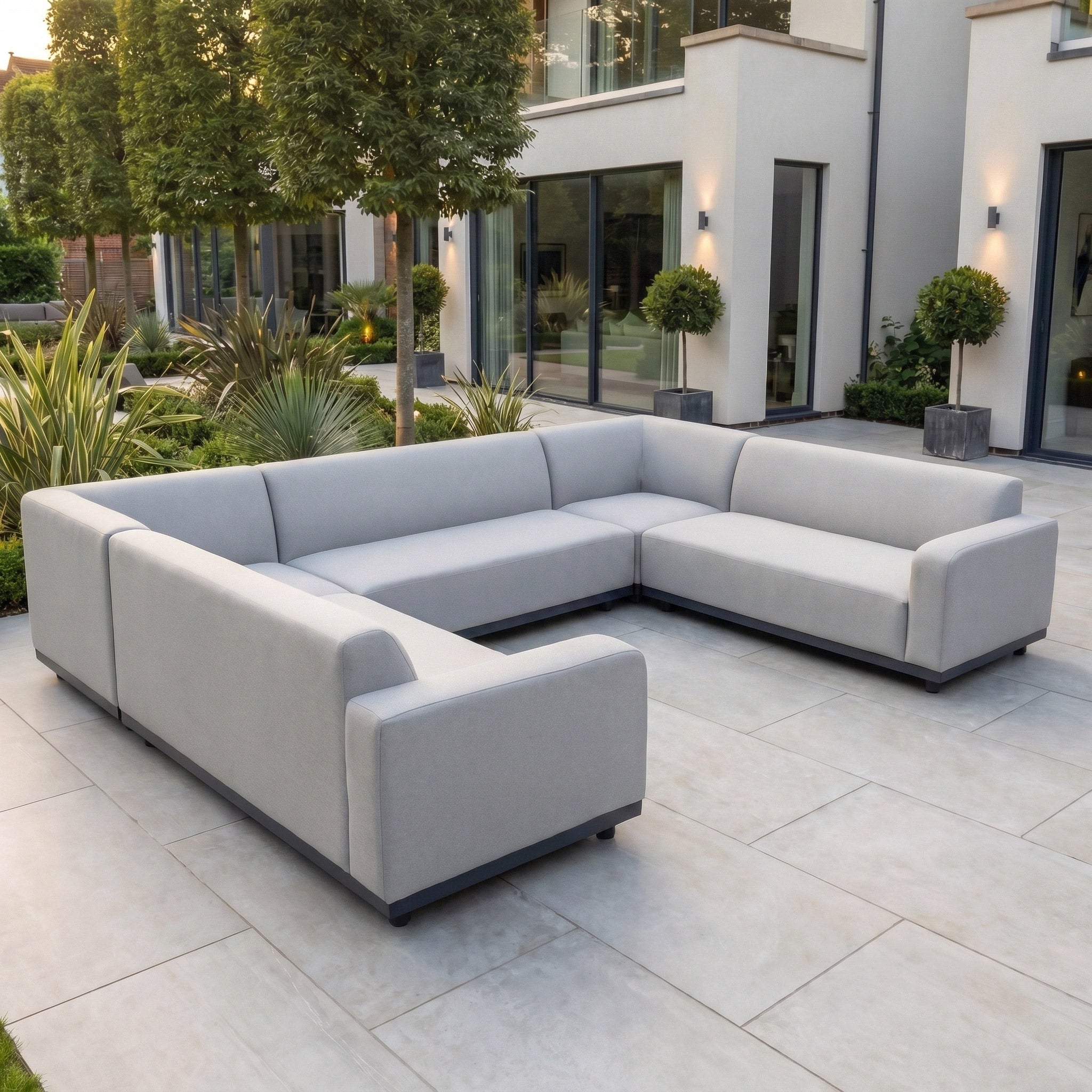 A Luna U-Shape Outdoor Fabric Sofa Set in Oyster Grey sits on a tiled patio outside a contemporary house, surrounded by greenery.